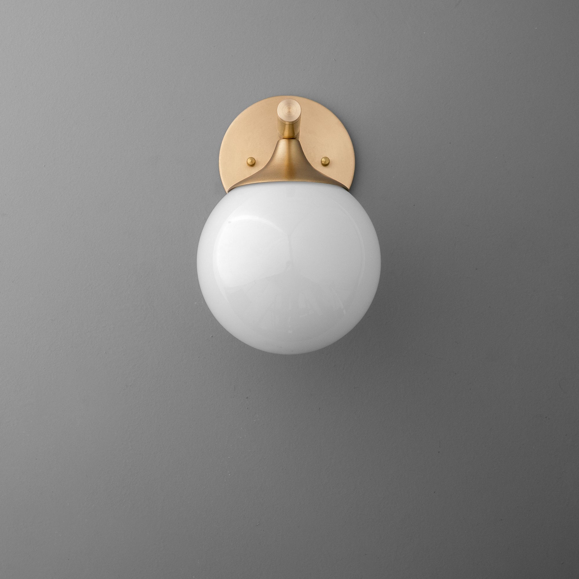 Wall sconce with round white glass globe and smooth raw brass mount on gray wall. - SCONCE MODEL NO. 1098
