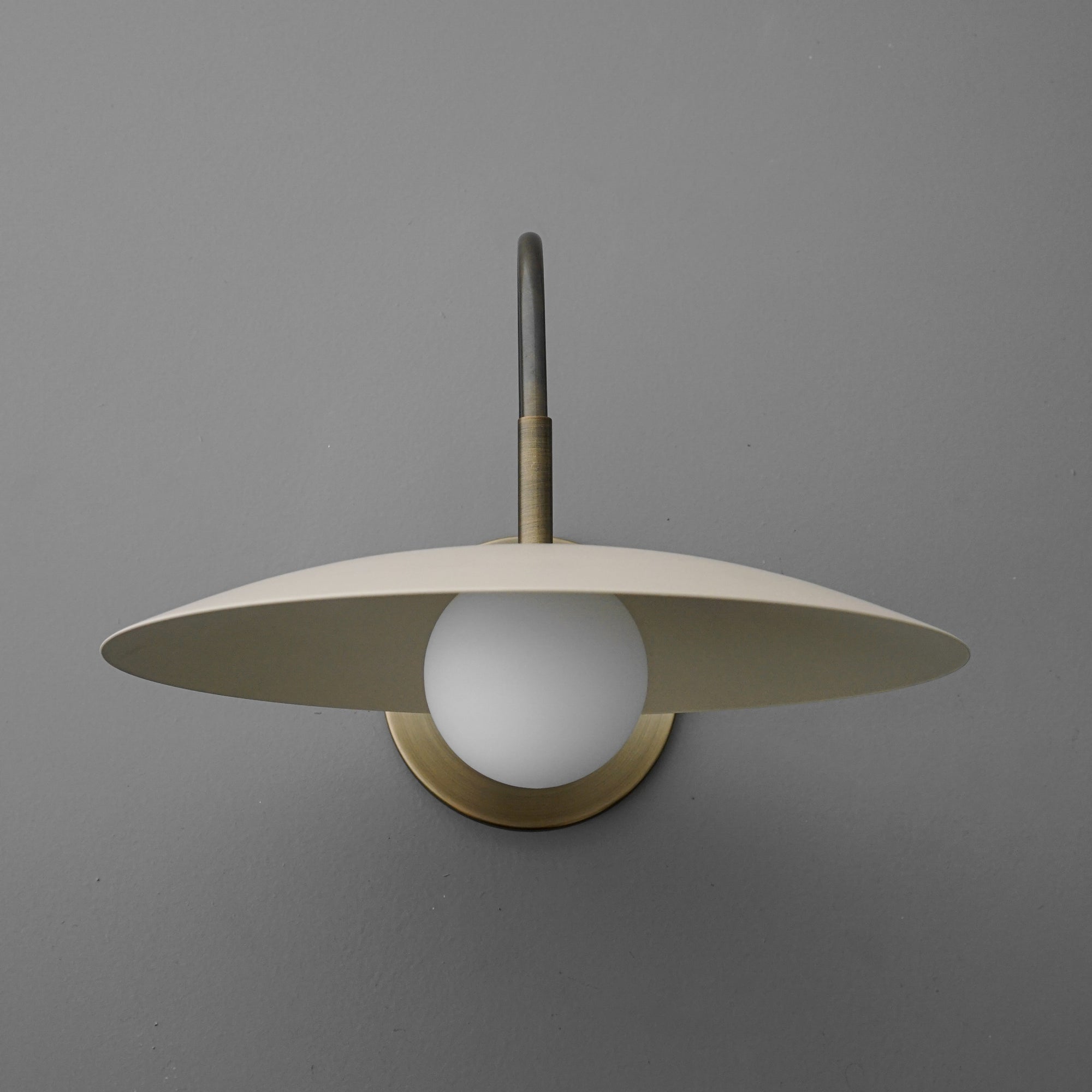 Antique brass wall sconce with a cream flat disc shade and white globe bulb against gray wall. - SCONCE MODEL NO. 9171