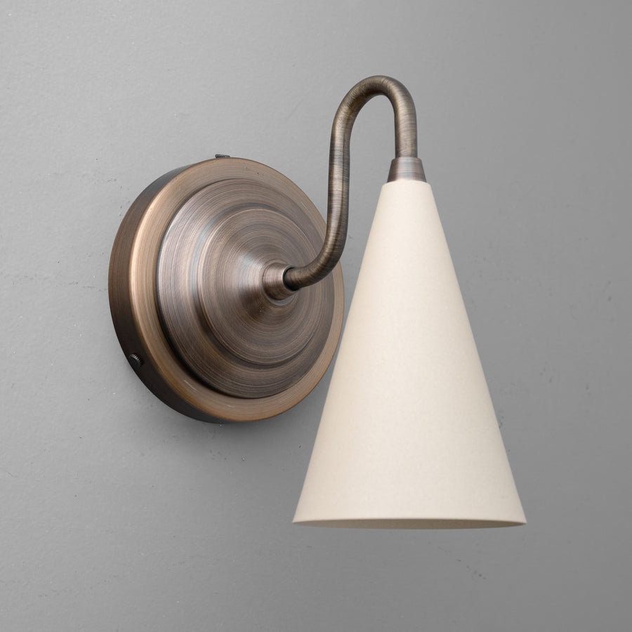 Antique brass sconce with curved arm and cream-colored conical fabric shade against gray wall. - Sconce Model No. 6790