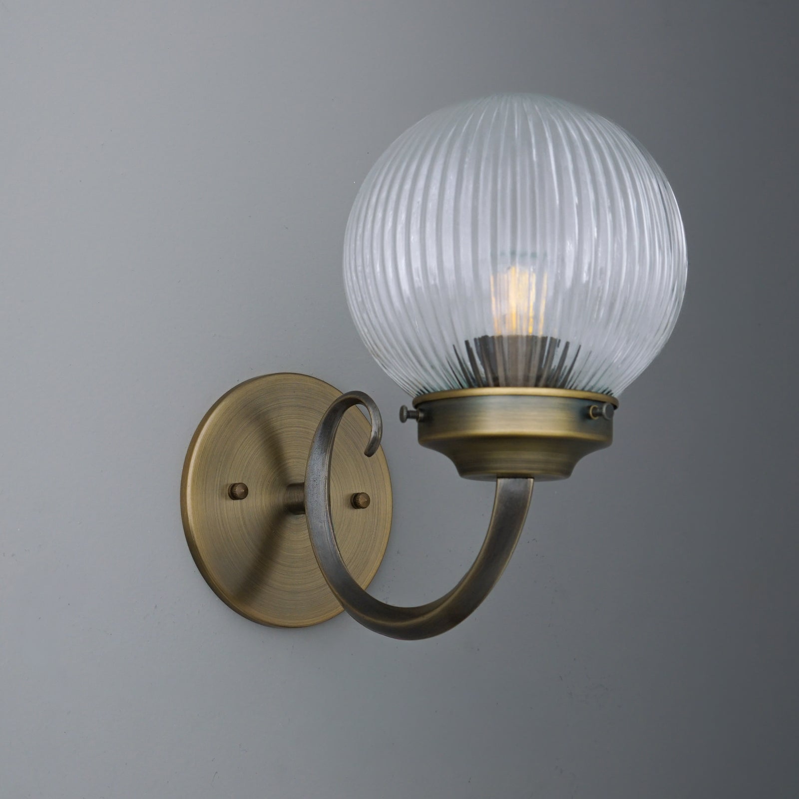 Wall sconce with antique brass curved arm and ribbed clear glass globe shade enclosing a single bulb. - SCONCE MODEL NO. 1093