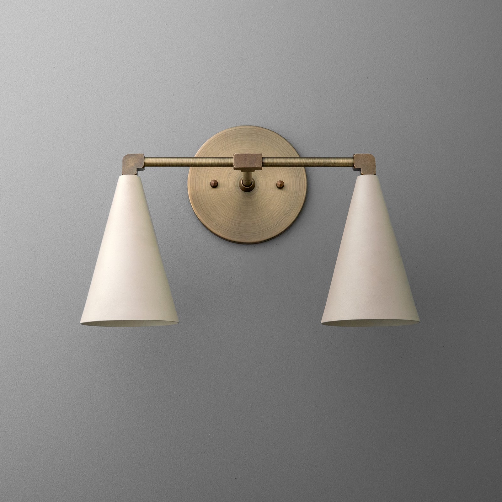 Two-light vanity light in Antique Brass with Cream cone shades on a straight arm and a round wall plate. - VANITY LIGHT MODEL NO. 1184