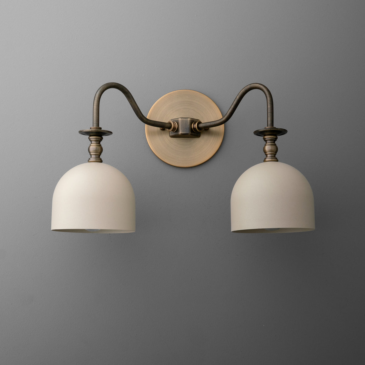 Two-light vanity light in Antique Brass with curved swan-neck arms and turned fitters holding steel bell shades, on a round wall plate. - VANITY LIGHT MODEL NO. 8710