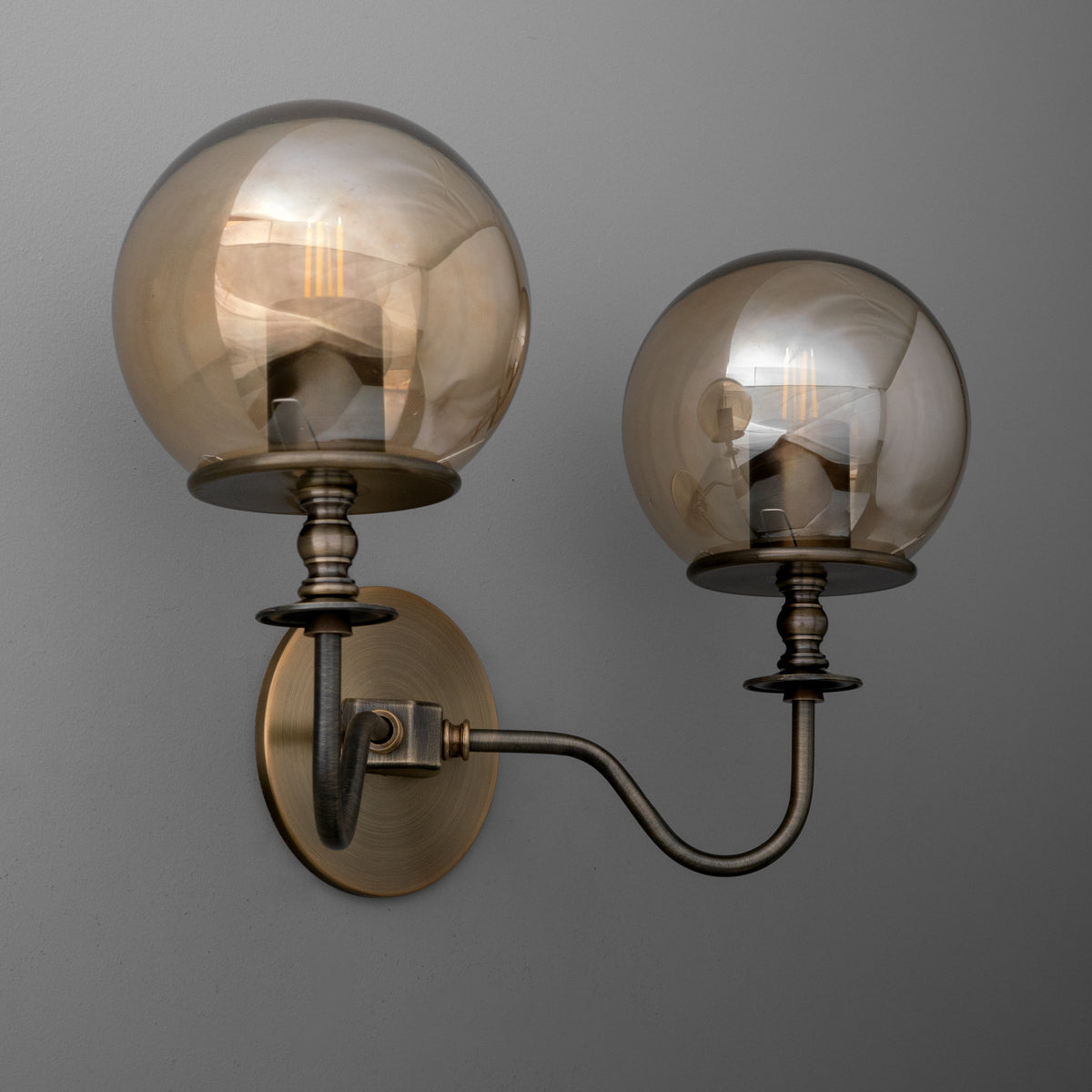 Angled view of a two-light sconce in Antique Brass with upright smoked glass globes over candle-style sockets on curved arms from a round wall plate. - SCONCE MODEL NO. 8251