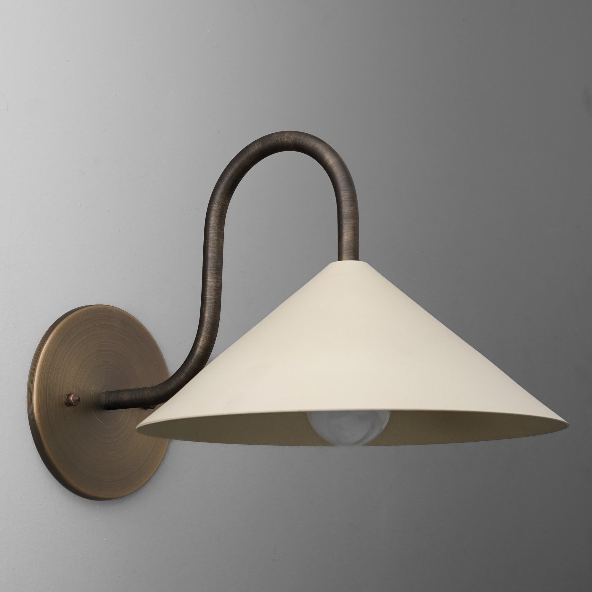 Wall-mounted sconce with a curved bronze arm and wide, cream-colored conical shade covering the bulb. - SCONCE MODEL NO. 1015