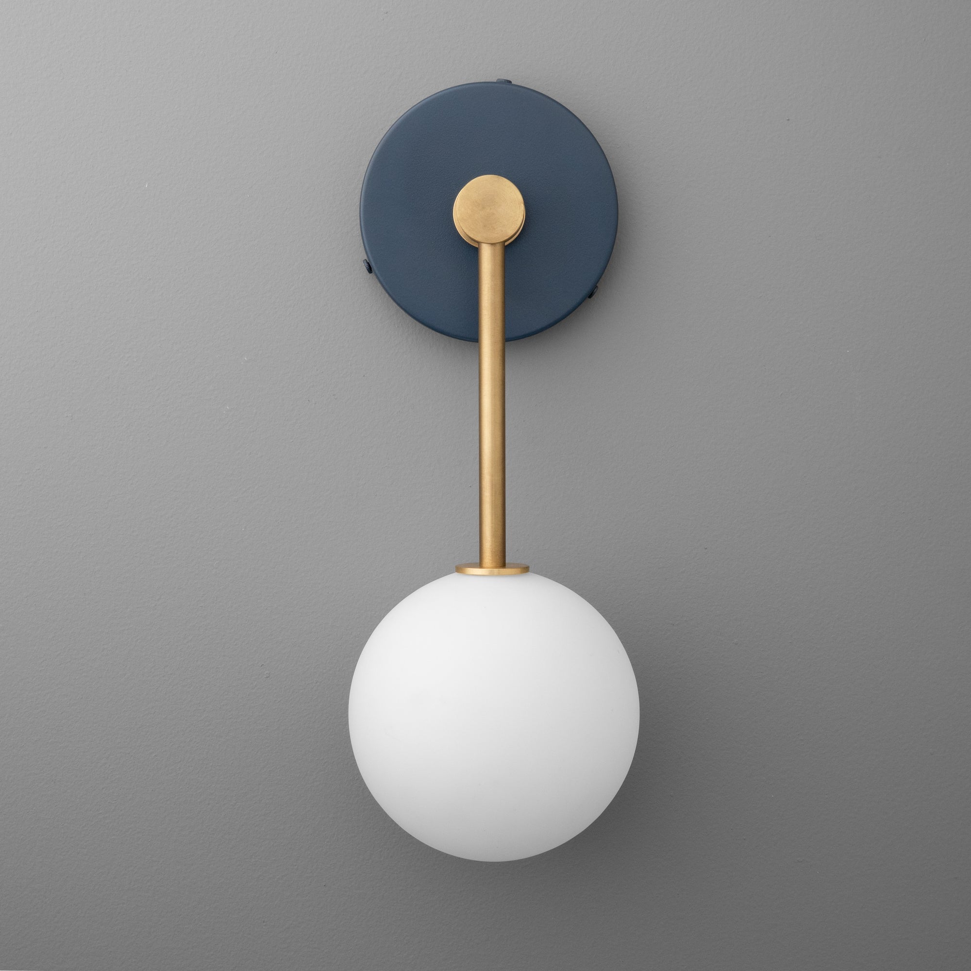 Raw brass wall sconce with coal blue circular mount and frosted white glass globe shade against gray background. - Sconce Model No. 5999