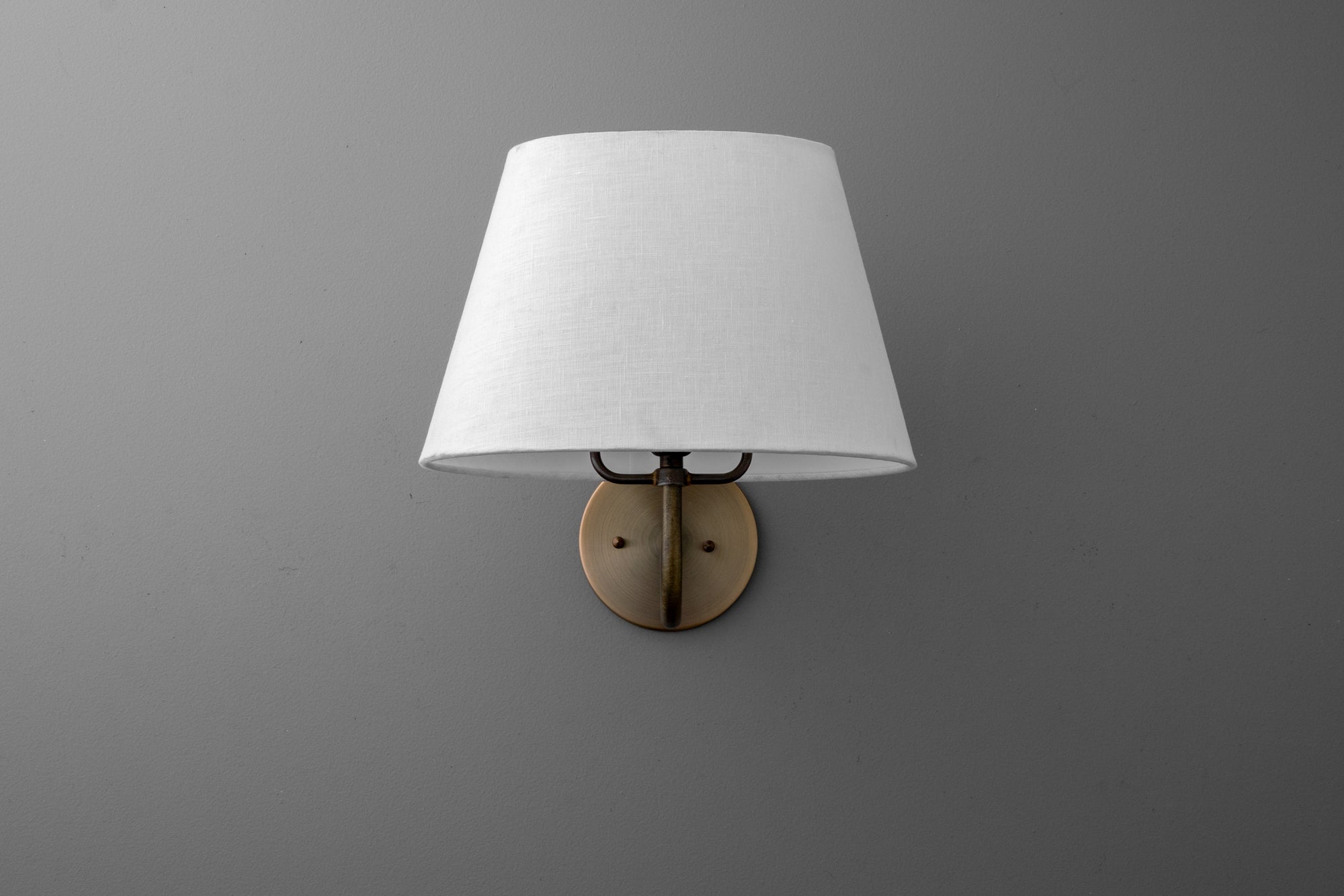 Wall sconce with round antique brass base and white fabric tapered shade against gray wall. - SCONCE MODEL NO. 0303