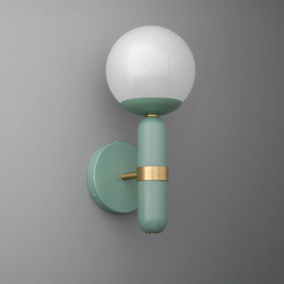 Wall sconce with round white glass globe and matte green cylindrical base with brass accent on gray wall. - SCONCE MODEL NO. 4786