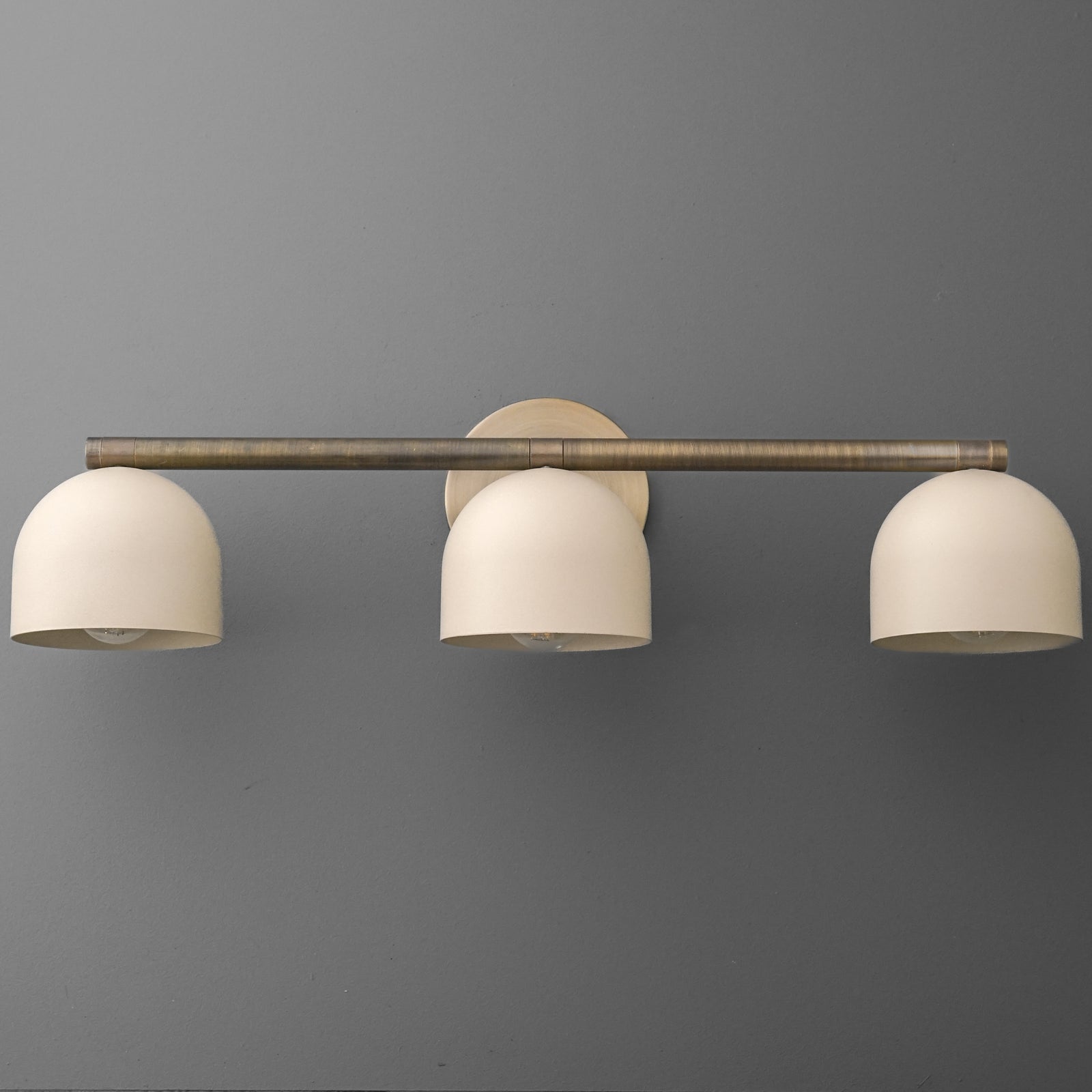 Three-light vanity light in Antique Brass with Cream dome shades hanging from a long straight bar on a round wall plate. - VANITY LIGHT MODEL NO. 1169
