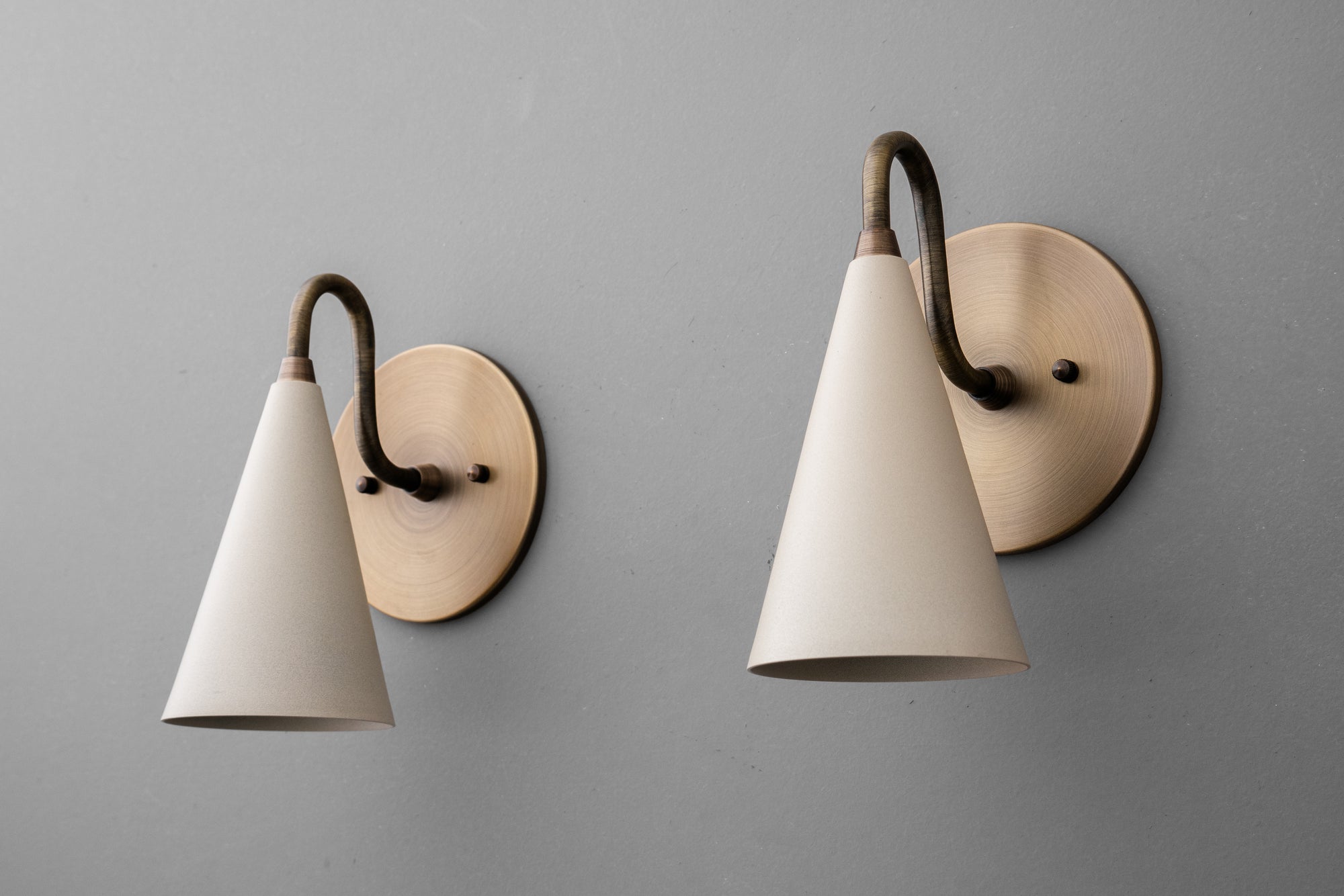Two wall sconces with antique brass finish and cream tapered conical shades on a gray wall. - Sconce Model No. 2900