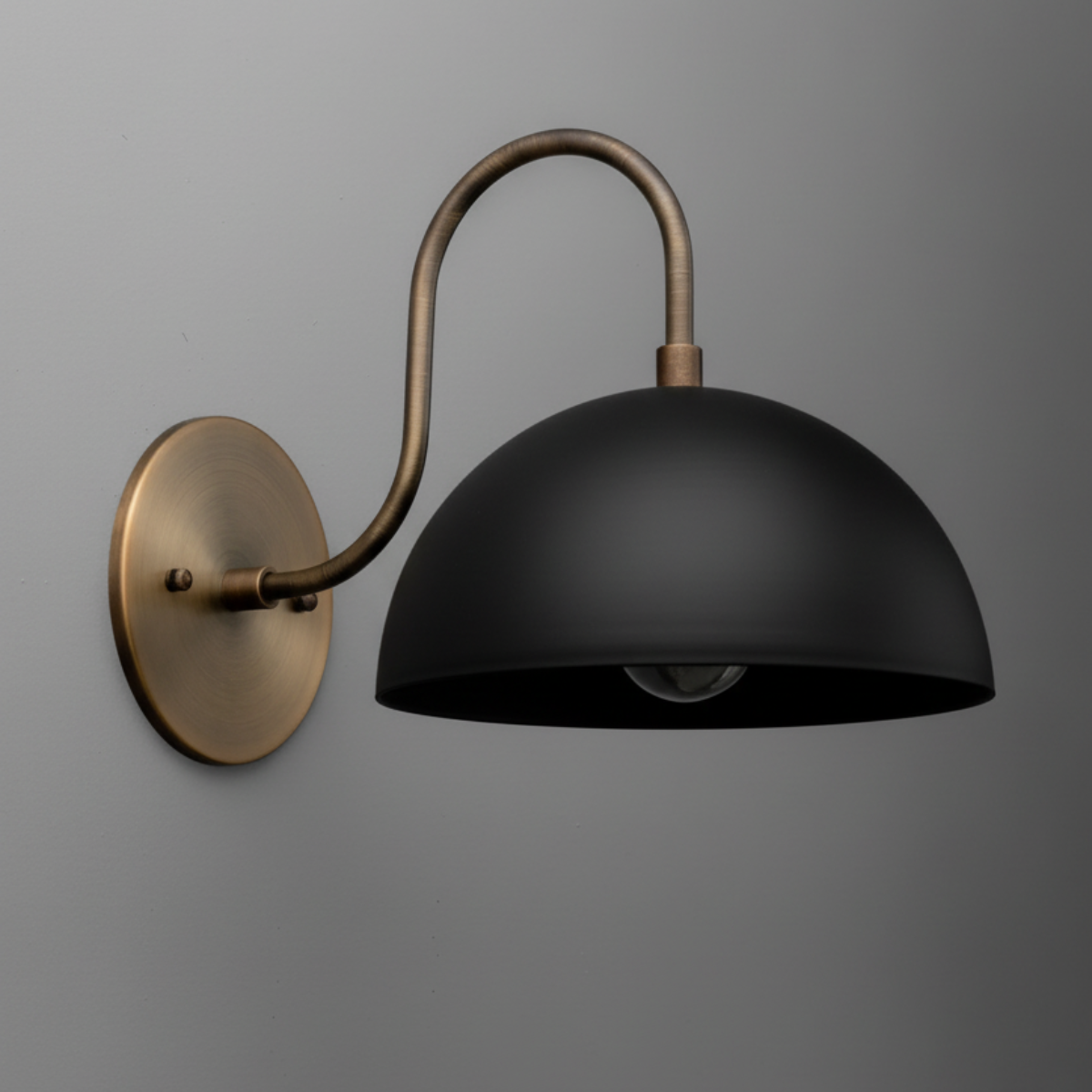 Matte black dome sconce with antique brass curved arm and round wall mount against gray wall. - SCONCE MODEL NO. 9092