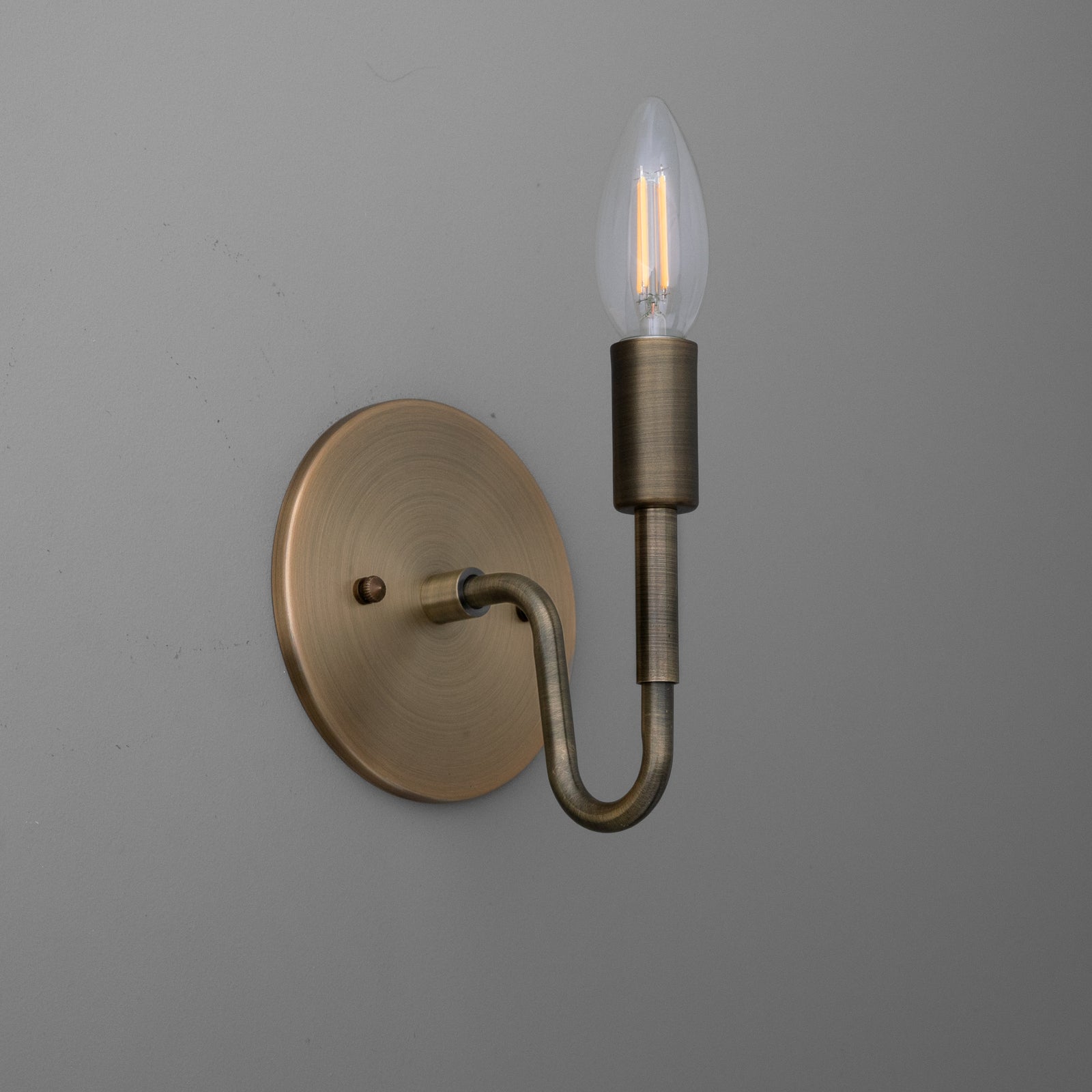 Antique brass wall sconce with a single curved arm and exposed candle-shaped bulb. - SCONCE MODEL NO. 1100