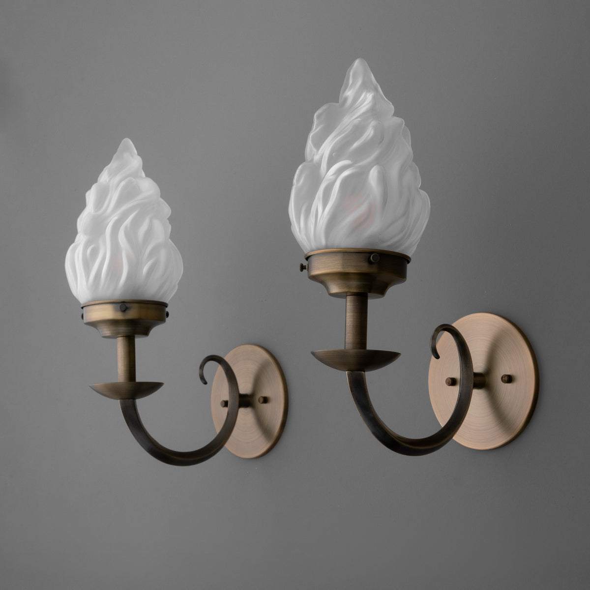 Pair of wall sconces with antique brass finish, each featuring a curved arm and frosted white flame-shaped glass shade. - SCONCE MODEL NO. 1057