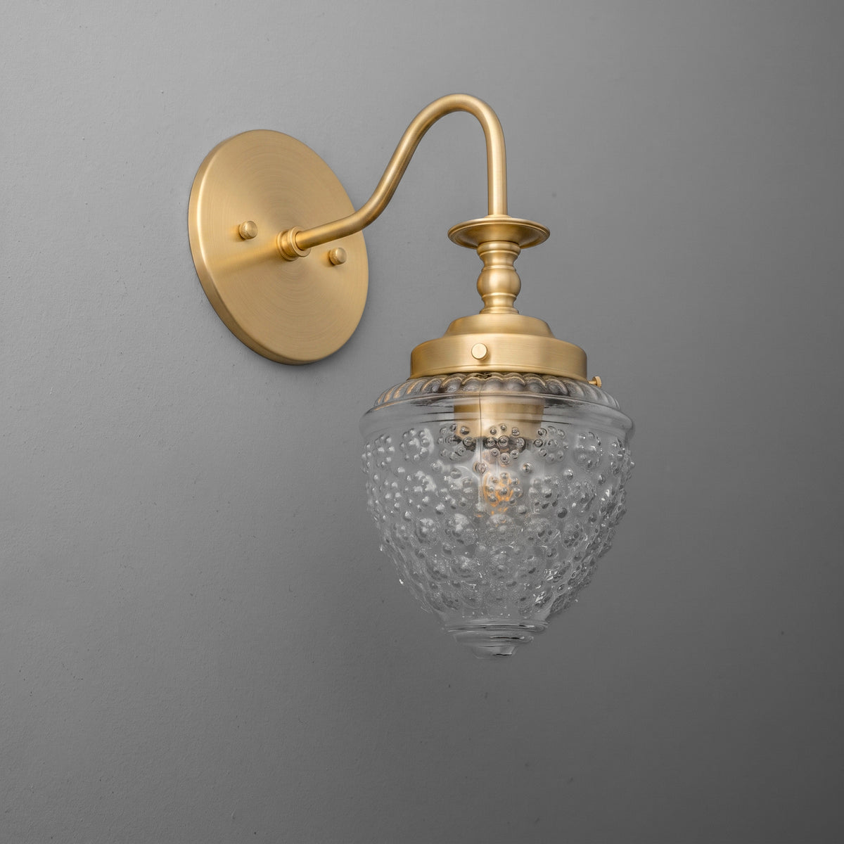 Matte gold metal wall sconce with curved arm and textured clear glass shade featuring bubble details. - SCONCE MODEL NO. 1138