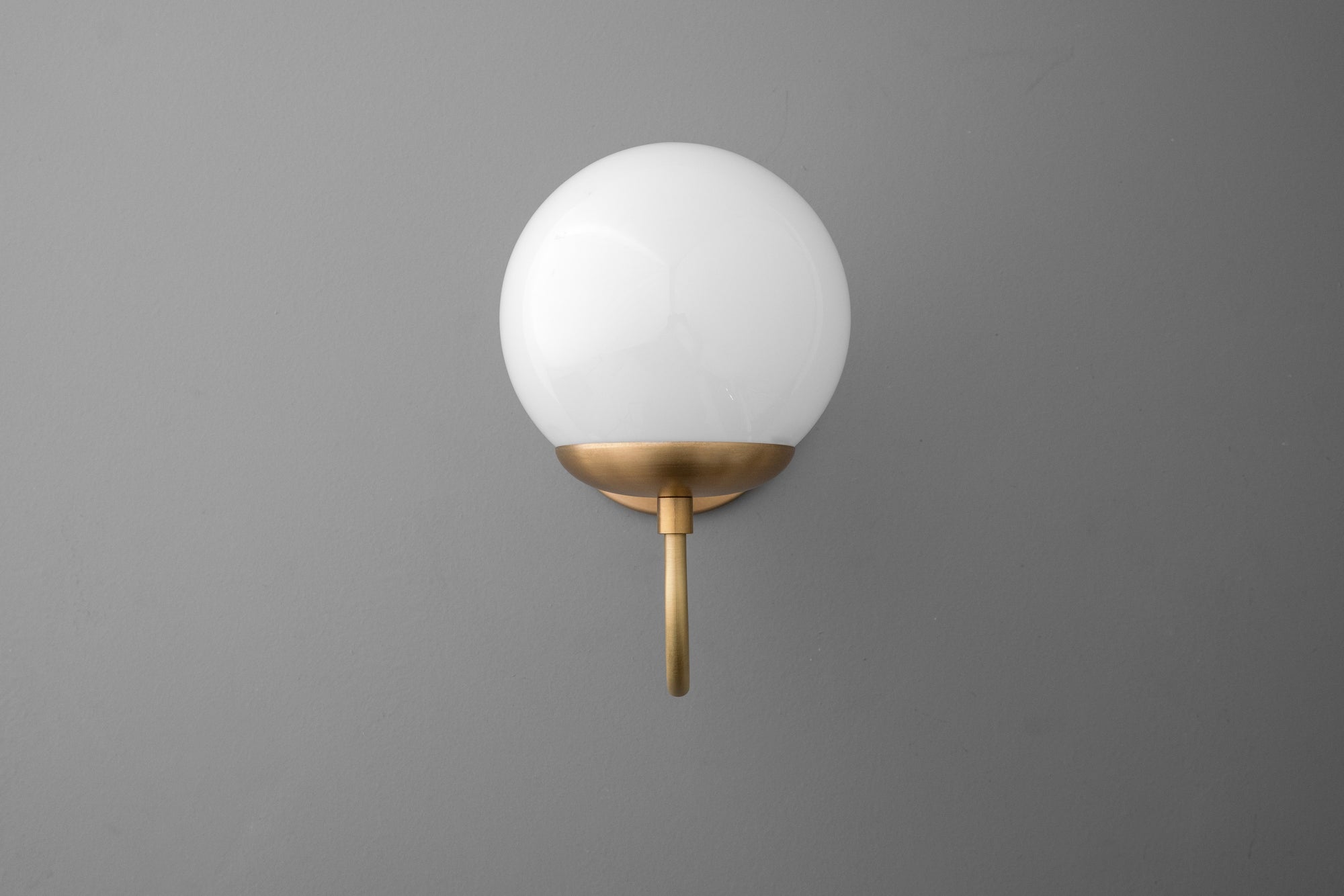 Wall sconce with a round white glass globe and raw brass mount on matte gray wall. - SCONCE MODEL NO. 3849