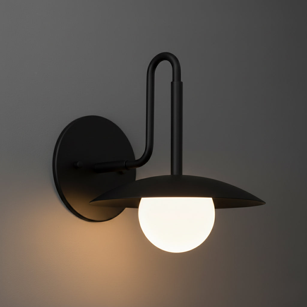 Black metal wall sconce with round backplate, upward curved arm, and glowing white globe bulb under flat circular shade. - SCONCE MODEL NO. 1017