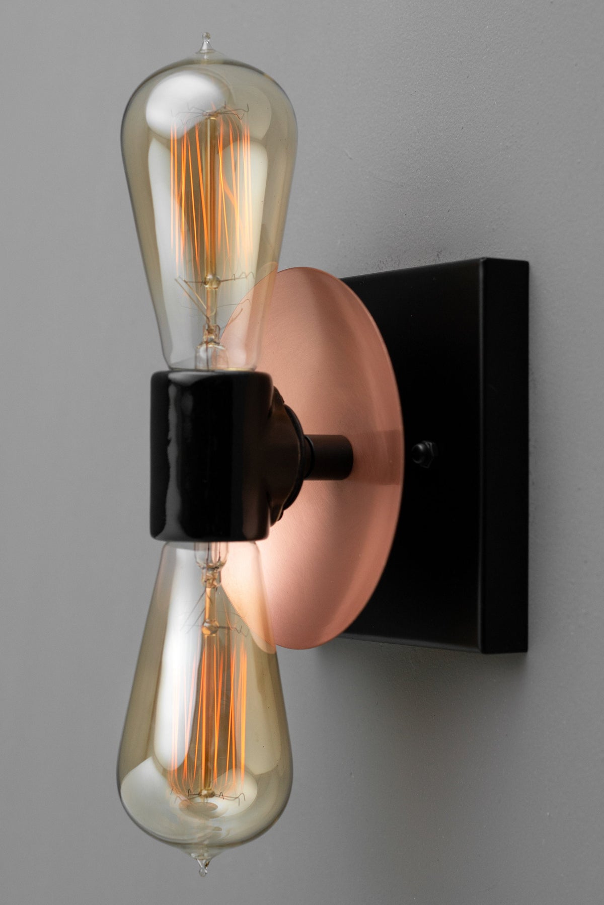 Industrial wall sconce with black fixture, copper round backplate, and dual exposed filament bulbs arranged vertically. - SCONCE MODEL NO. 8169