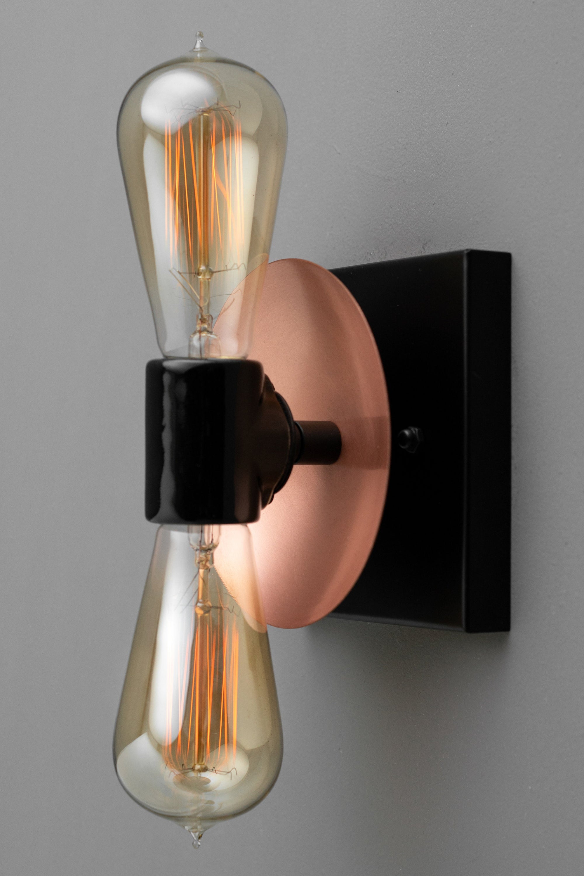 Industrial wall sconce with black fixture, copper round backplate, and dual exposed filament bulbs arranged vertically. - SCONCE MODEL NO. 8169