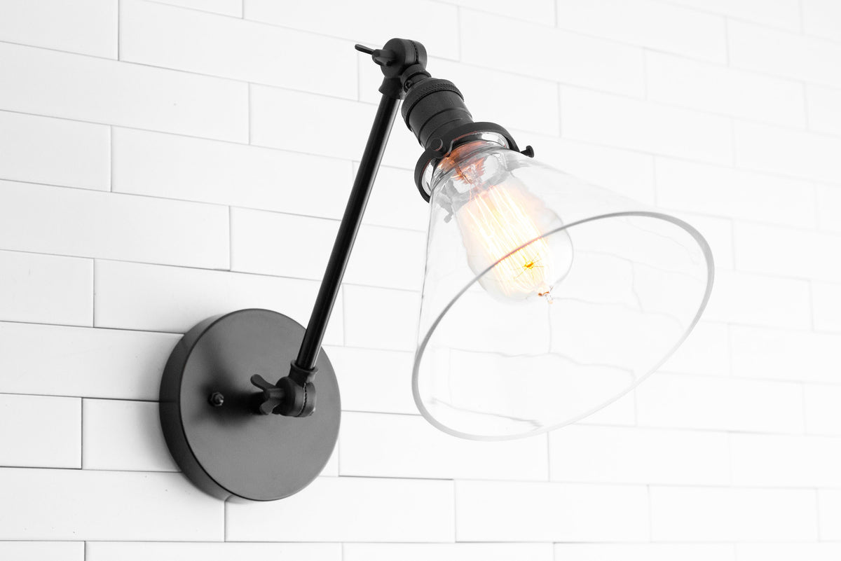 Black adjustable industrial wall sconce with clear glass shade and exposed bulb on white tile wall. - SCONCE MODEL NO. 7962