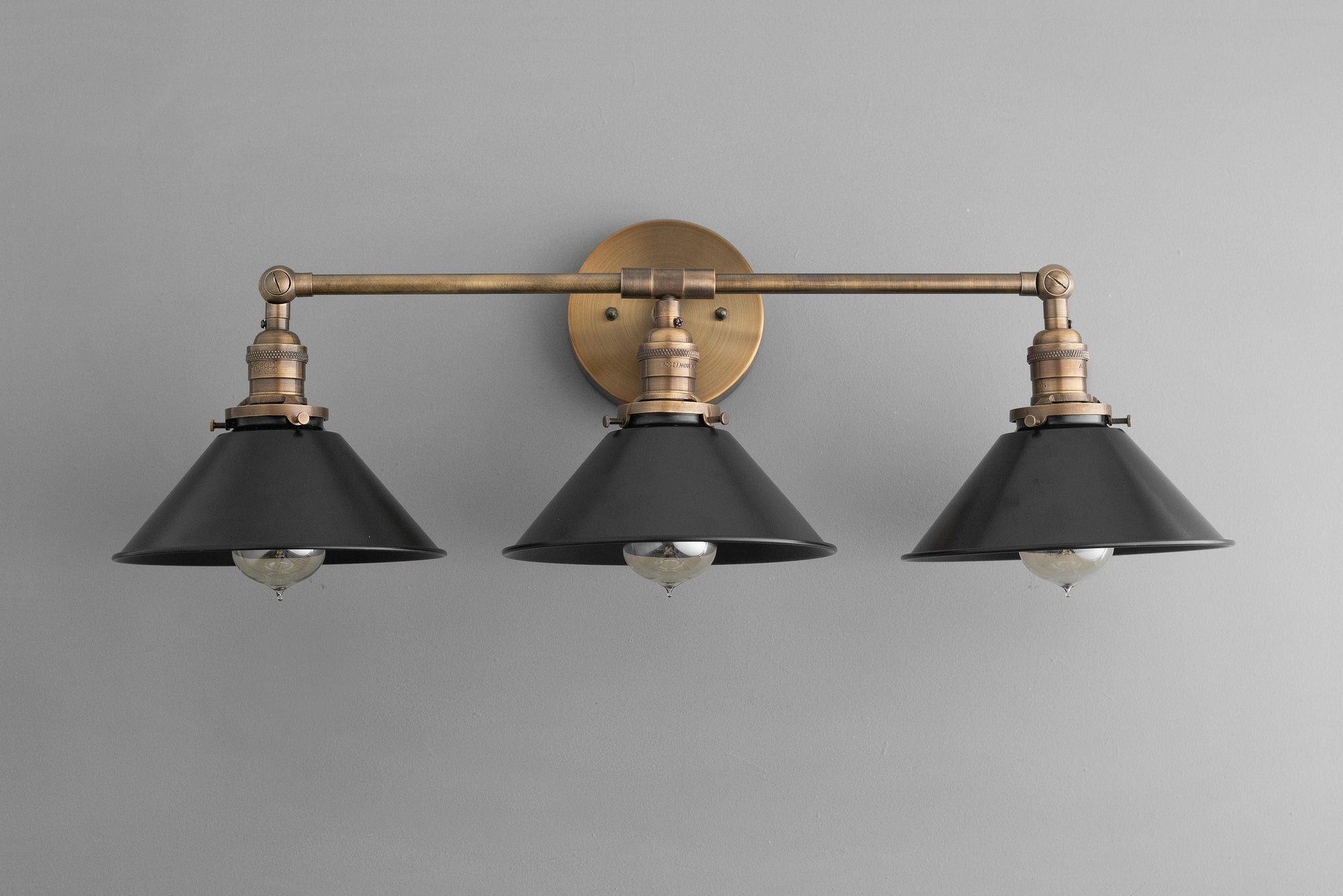 Three-bulb industrial vanity light with antique brass finish and black metal shades mounted on a round wall plate. - VANITY LIGHT MODEL NO. 3406