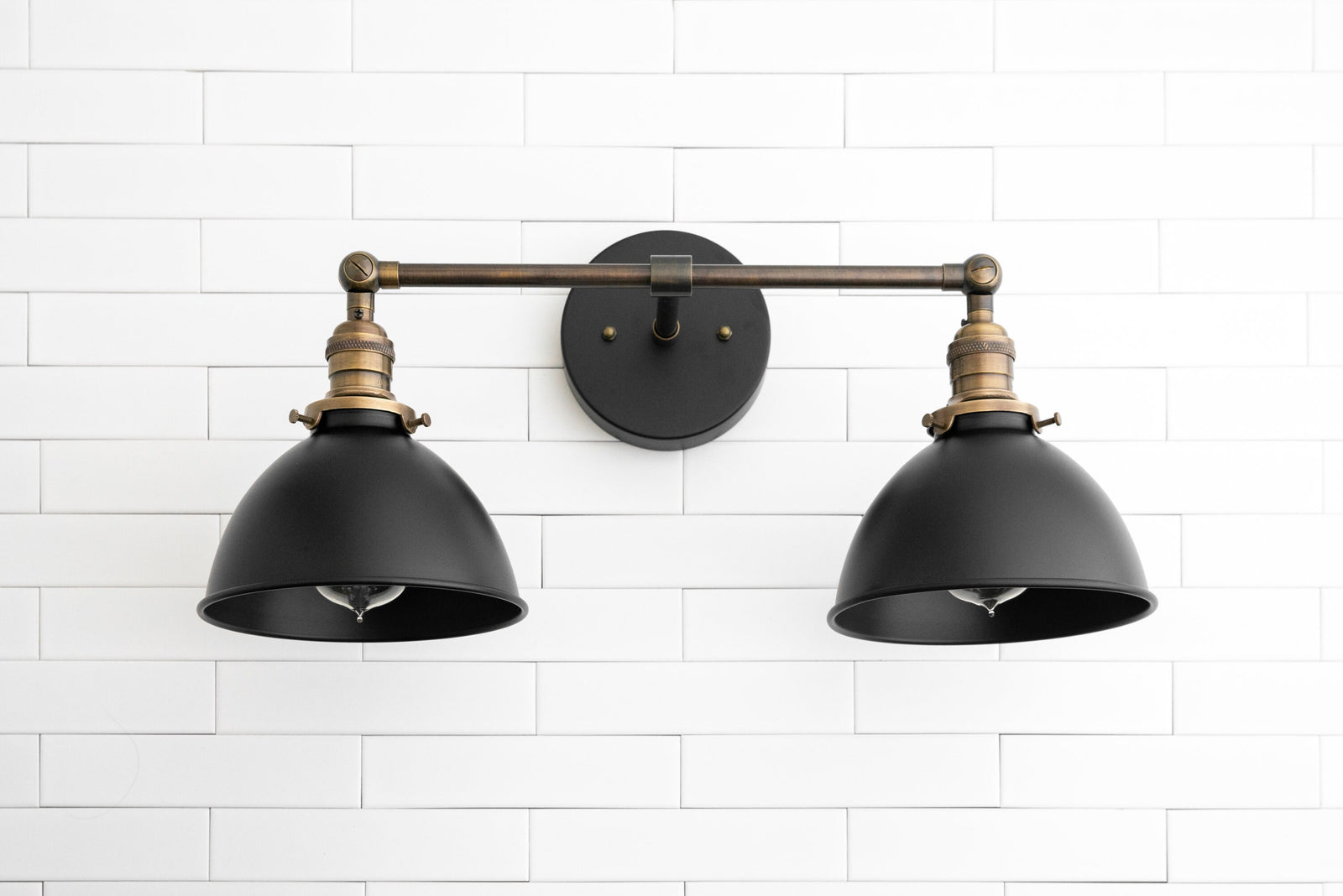 Two-light industrial vanity sconce with black shades and antique brass arms on white tile wall. - SCONCE MODEL NO. 8791