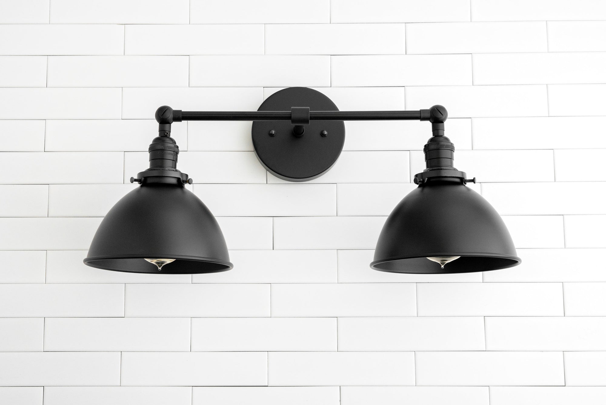 Black industrial 2-light vanity sconce with adjustable arms and dome shades on white subway tile wall. - SCONCE MODEL NO. 8791