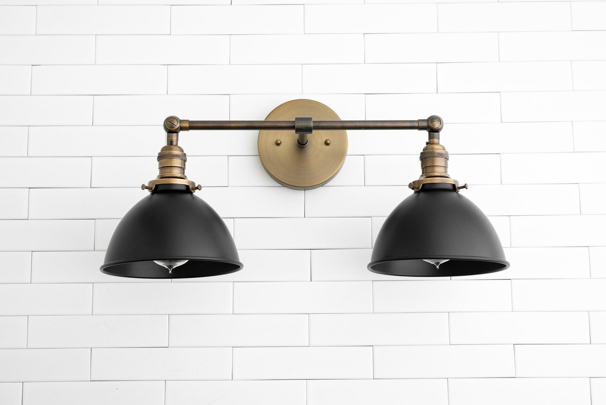 Industrial style 2-light vanity sconce with black shades and antique brass fixture mounted on white tile wall. - SCONCE MODEL NO. 8791