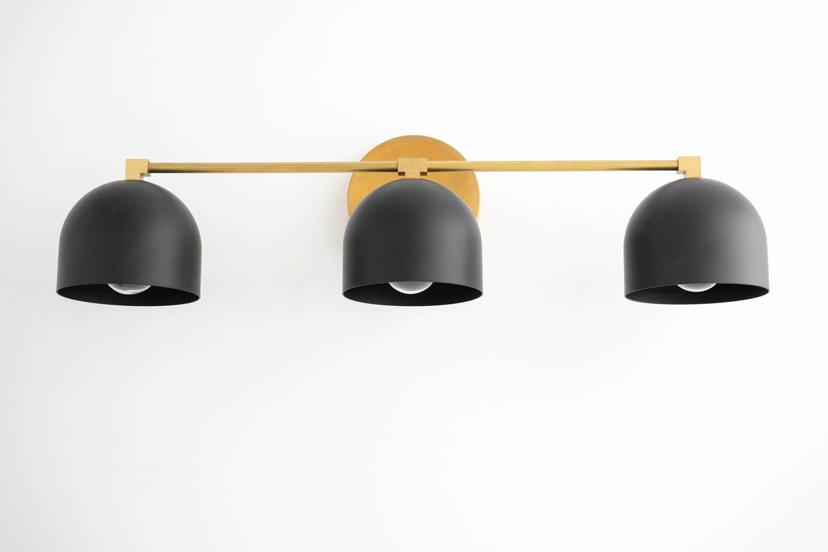 Three matte black dome shades mounted on a horizontal antique brass bar vanity light with round wall plate. - VANITY LIGHT MODEL NO. 7565
