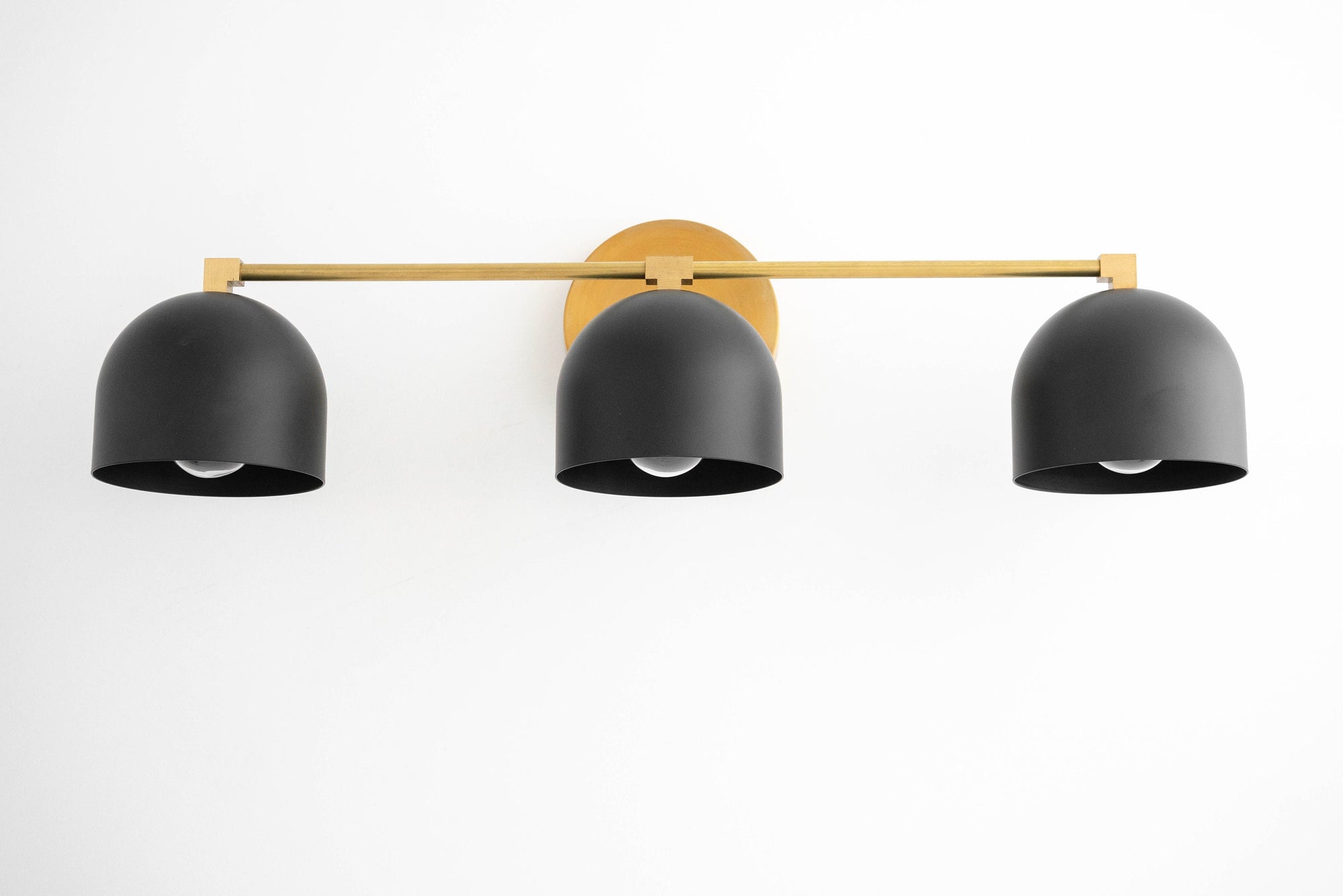 Three matte black dome shades mounted on a horizontal antique brass bar vanity light with round wall plate. - VANITY LIGHT MODEL NO. 7565