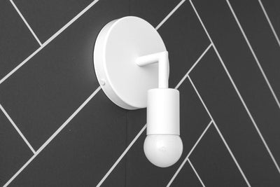 White metal wall sconce with exposed bulb and round base on dark gray tiled wall. - SCONCE MODEL NO. 5480