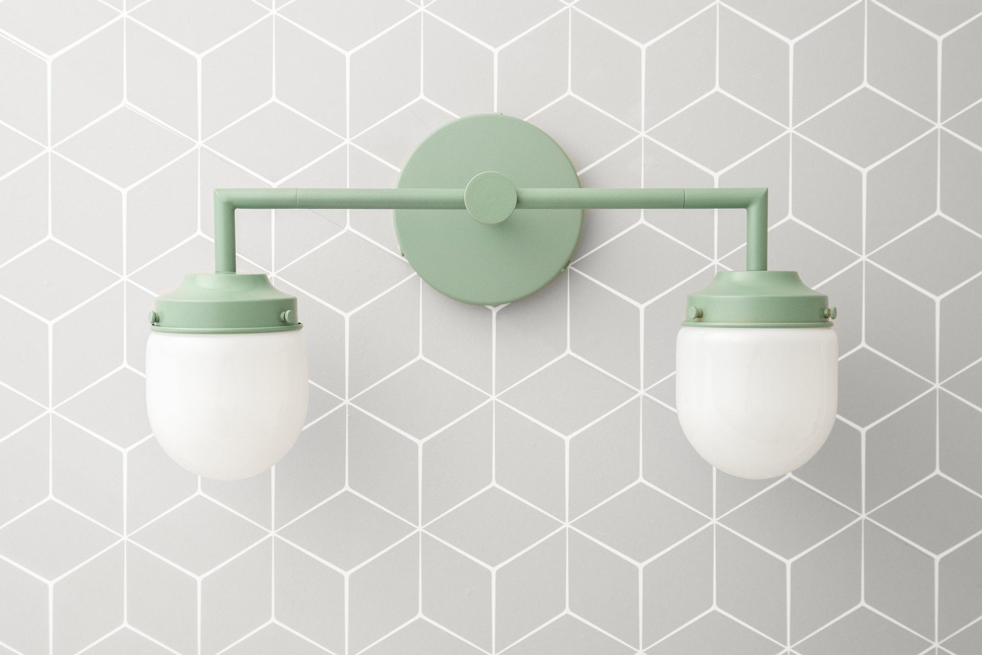 Matte sage green art deco vanity light with two frosted white glass globe shades mounted on geometric gray tile background. - VANITY LIGHT MODEL NO. 7536