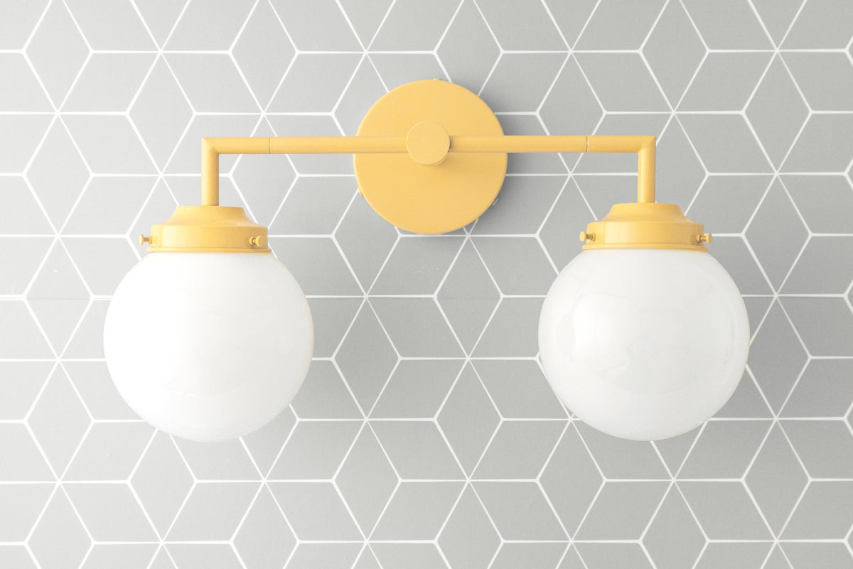 Yellow metal vanity light with two round white glass globe bulbs mounted on geometric gray tile wall. - VANITY LIGHT MODEL NO. 9500