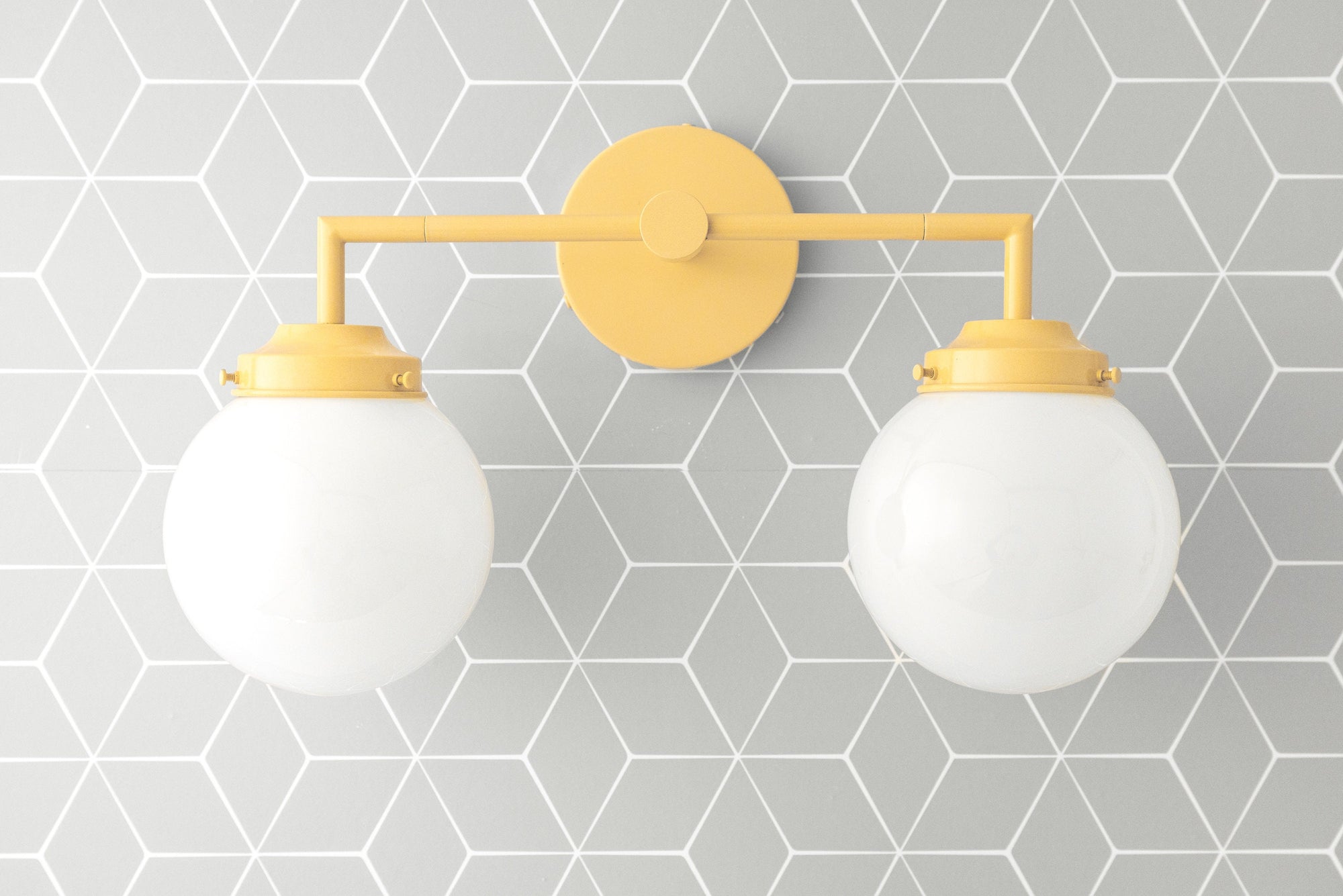 Yellow metal vanity light with two round white glass globe bulbs mounted on geometric gray tile wall. - VANITY LIGHT MODEL NO. 9500