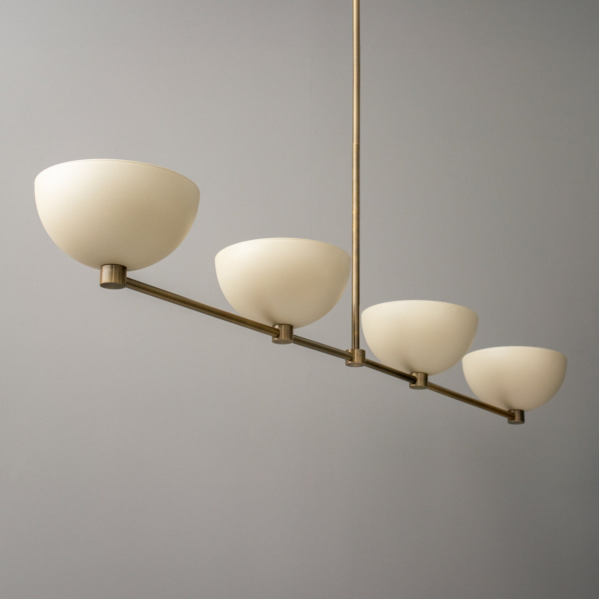 Antique brass linear chandelier with four cream dome-shaped glass shades against gray background. - CHANDELIER MODEL NO. 4577