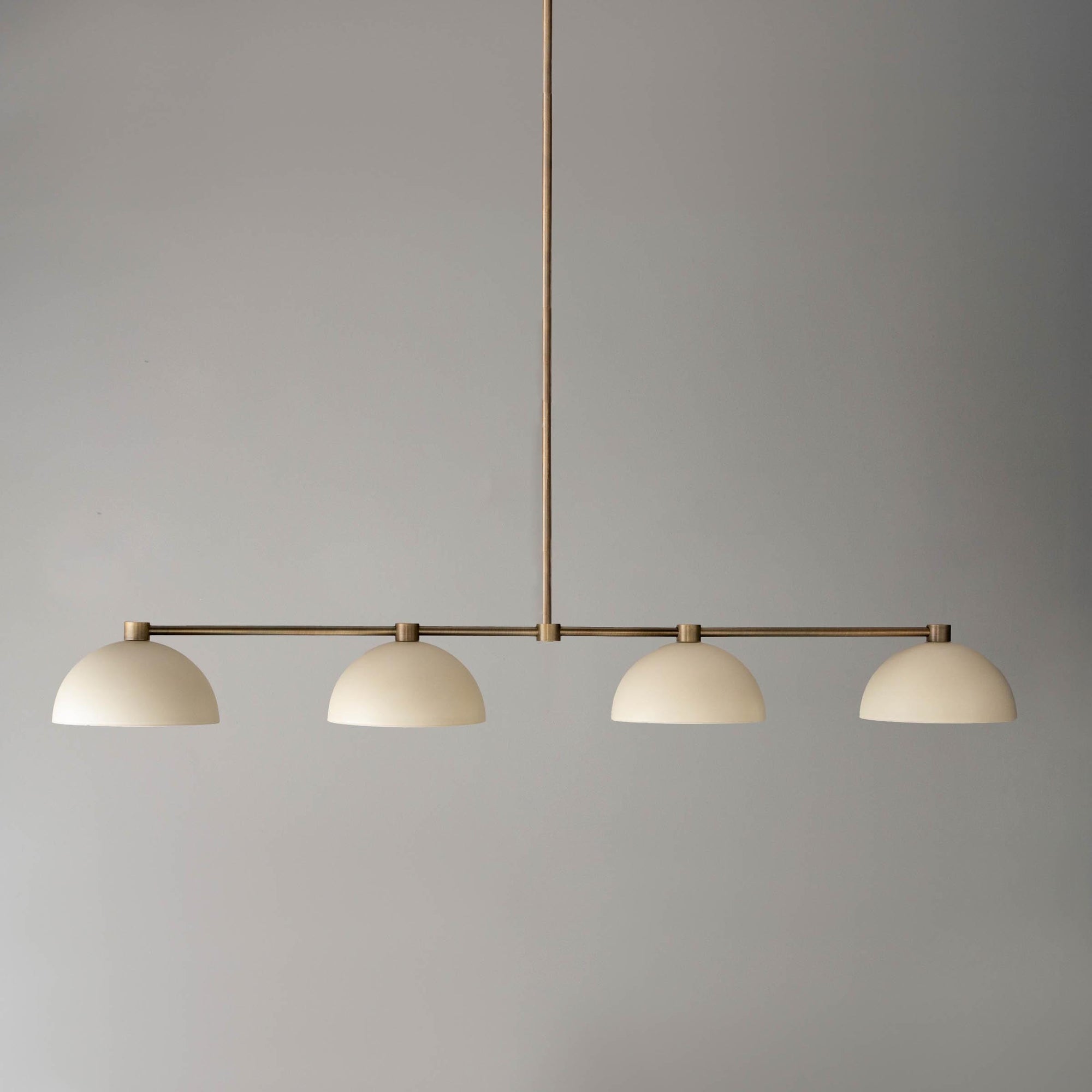 Antique brass linear chandelier with four cream dome-shaped shades hanging from a central rod. - CHANDELIER MODEL NO. 4577