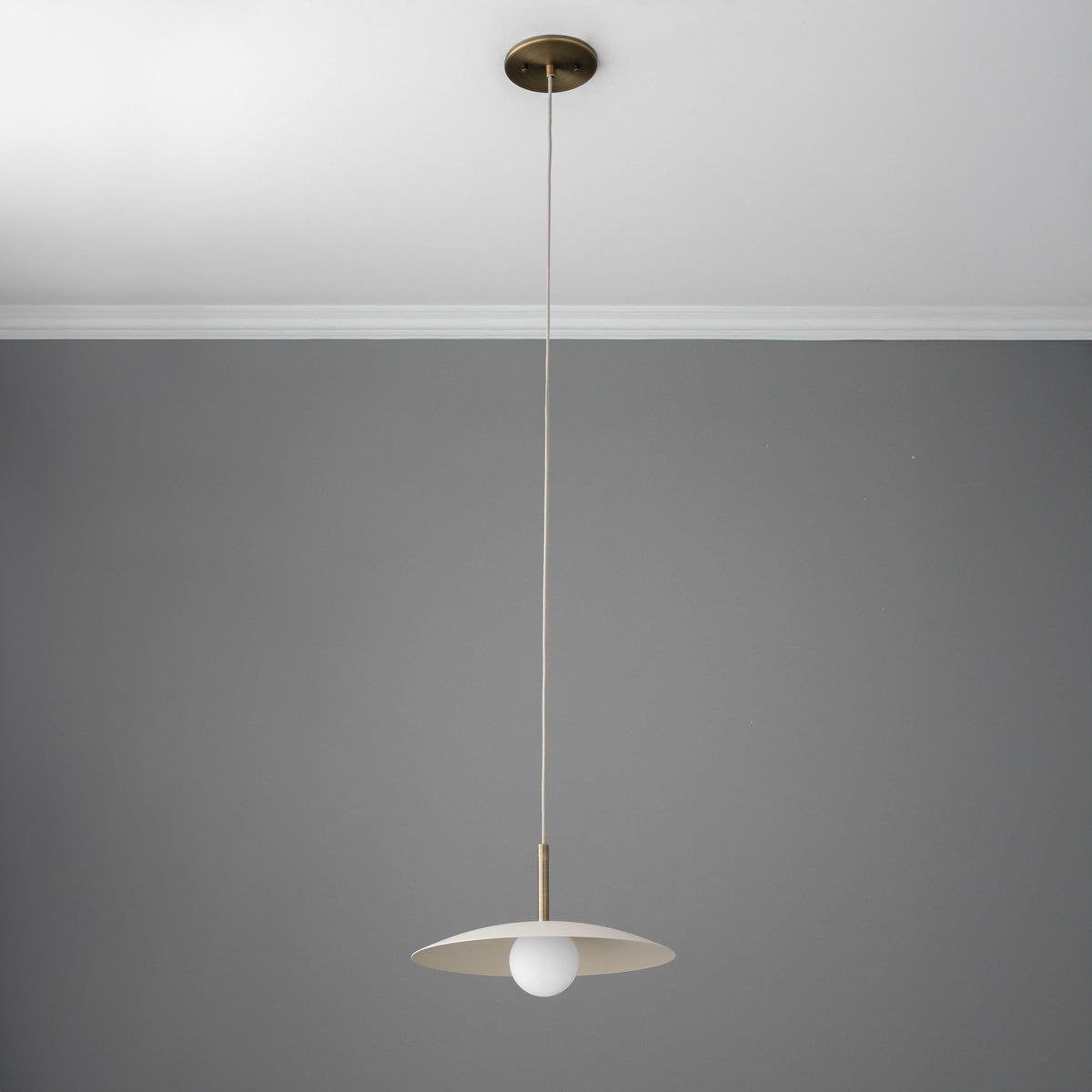 Minimalist pendant light with wide, flat antique brass shade and tan cord against gray ceiling and wall. - PENDANT MODEL NO. 5693