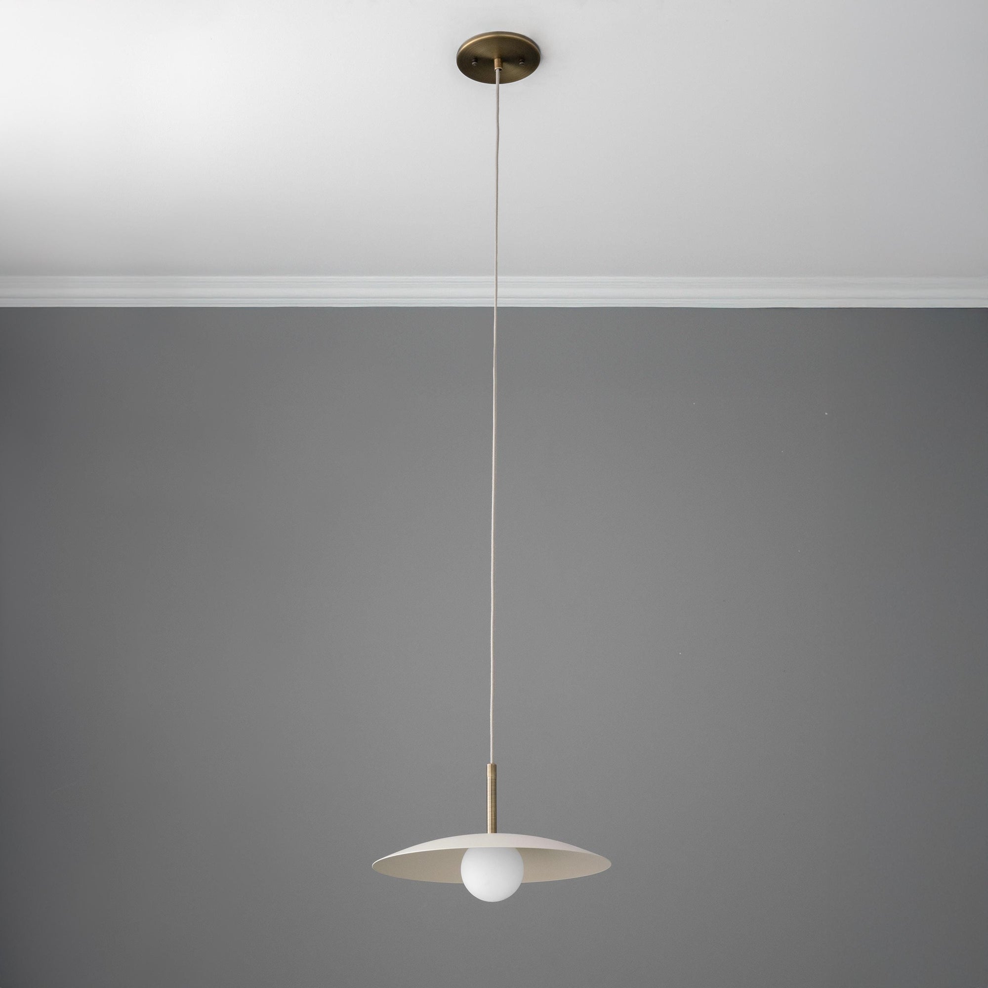 Minimalist pendant light with wide, flat antique brass shade and tan cord against gray ceiling and wall. - PENDANT MODEL NO. 5693