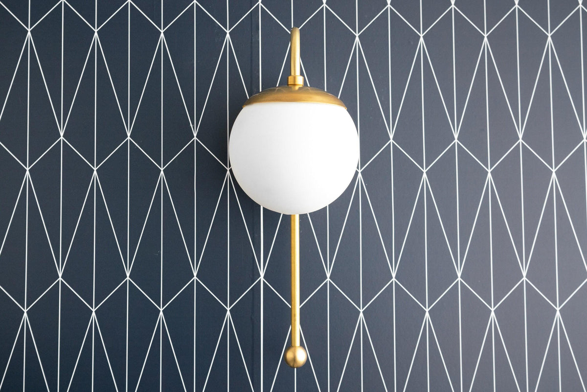 Art Deco style wall sconce with white globe shade and polished brass finish against geometric dark blue wallpaper. - SCONCE MODEL NO. 1045