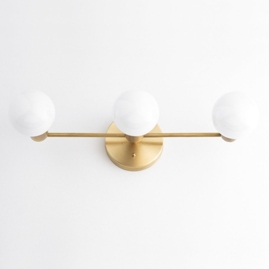 Mid-century modern vanity light with three white globe bulbs on a raw brass horizontal bar and round wall mount. - VANITY LIGHT MODEL NO. 0518
