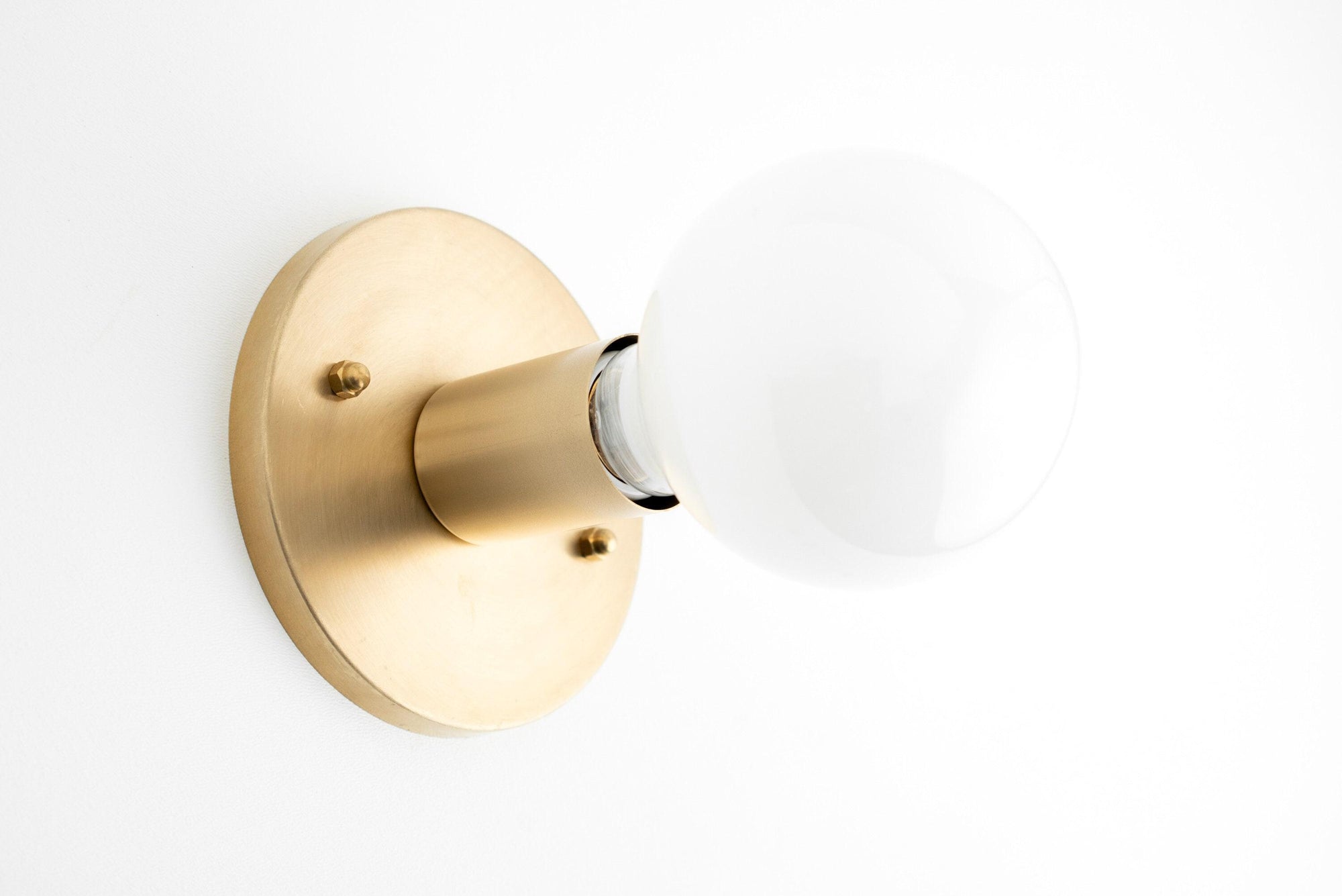 Mid-century modern raw brass wall sconce with exposed round white bulb and circular base. - SCONCE MODEL NO. 2057