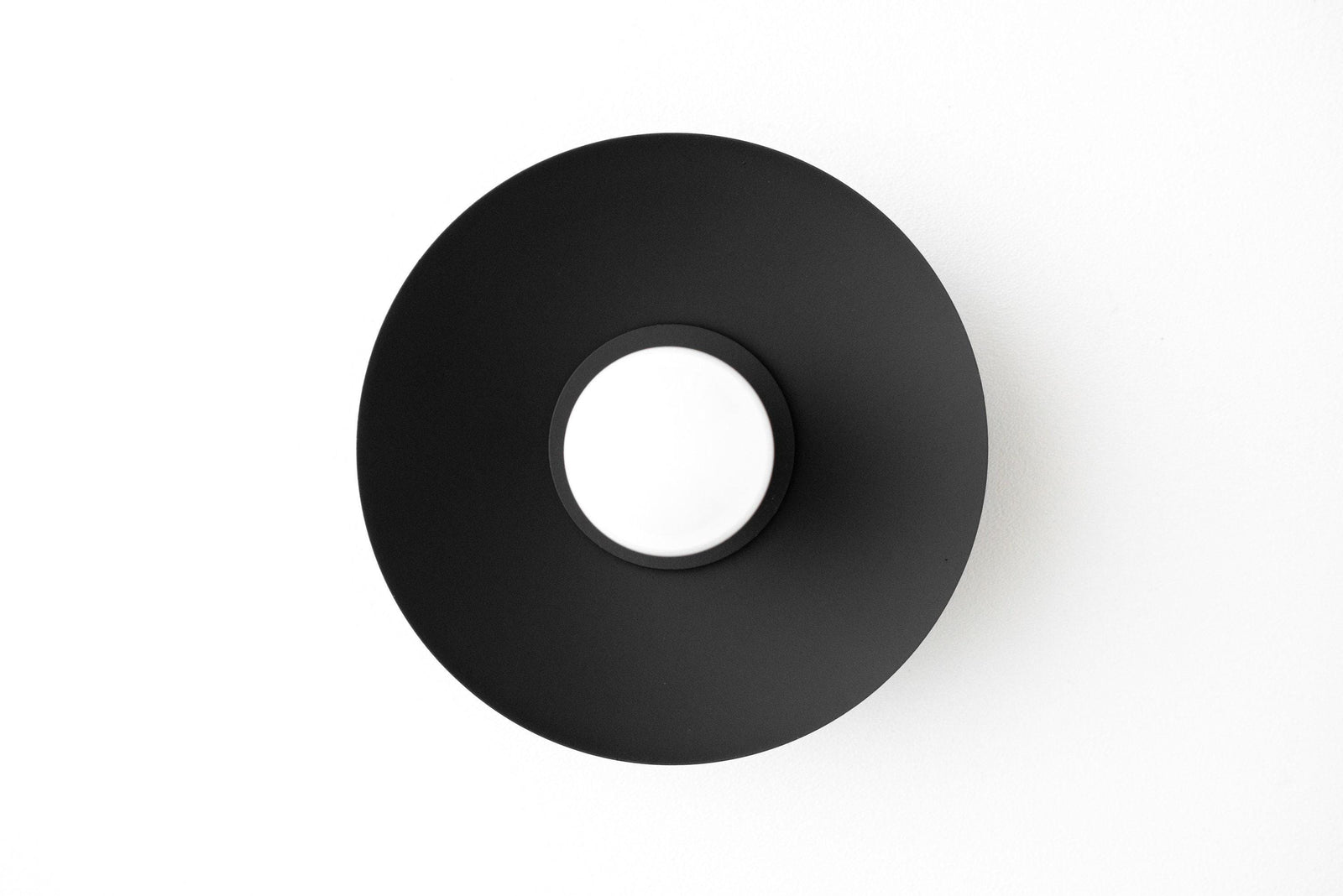 Black matte circular metal wall sconce with exposed bulb socket center and flat, wide shade. - SCONCE MODEL NO. 7004