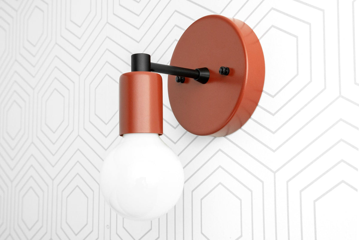 Scandinavian-style wall sconce in matte orange with exposed bulb and black arm on white geometric wall. - SCONCE MODEL NO. 9036
