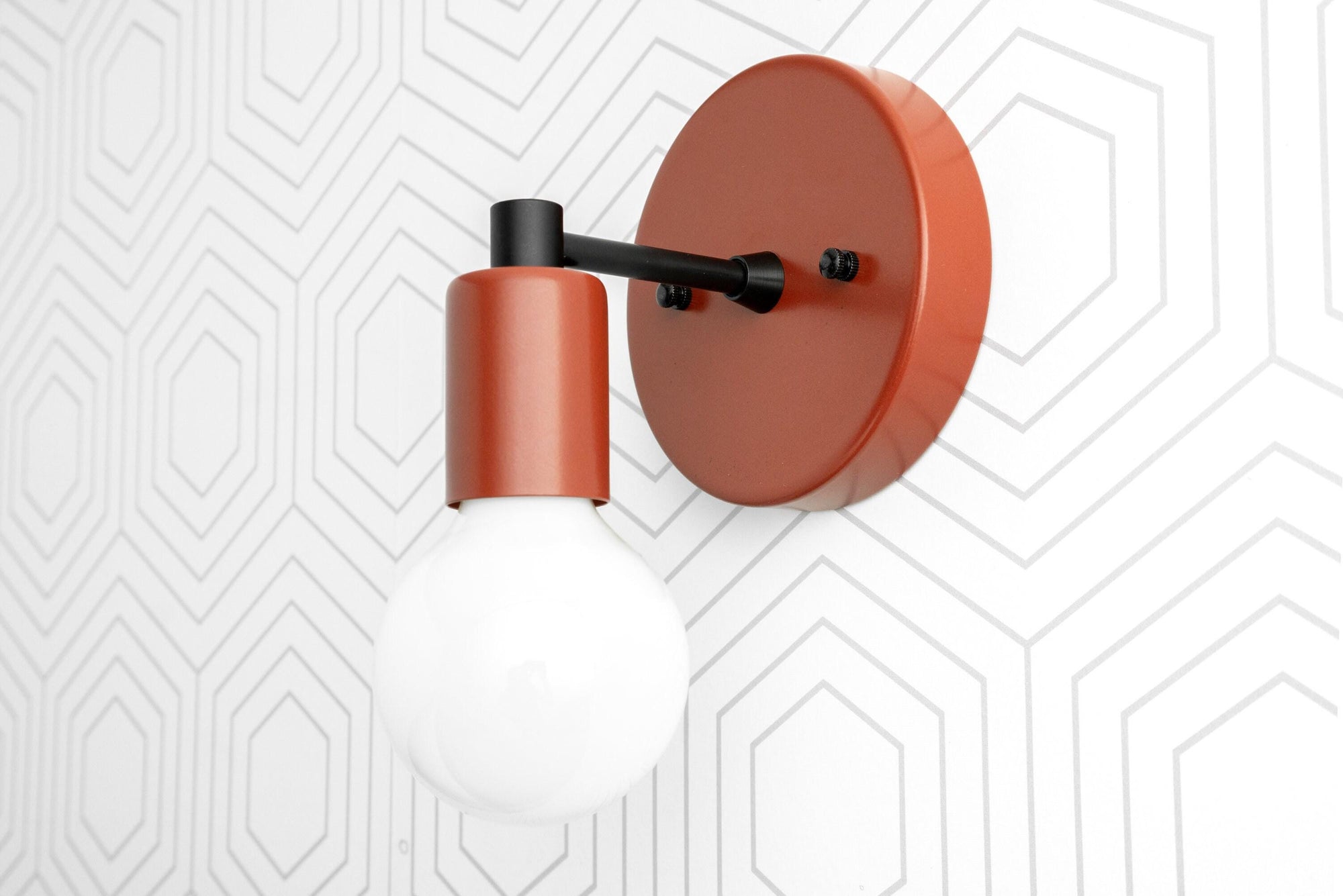 Scandinavian-style wall sconce in matte orange with exposed bulb and black arm on white geometric wall. - SCONCE MODEL NO. 9036