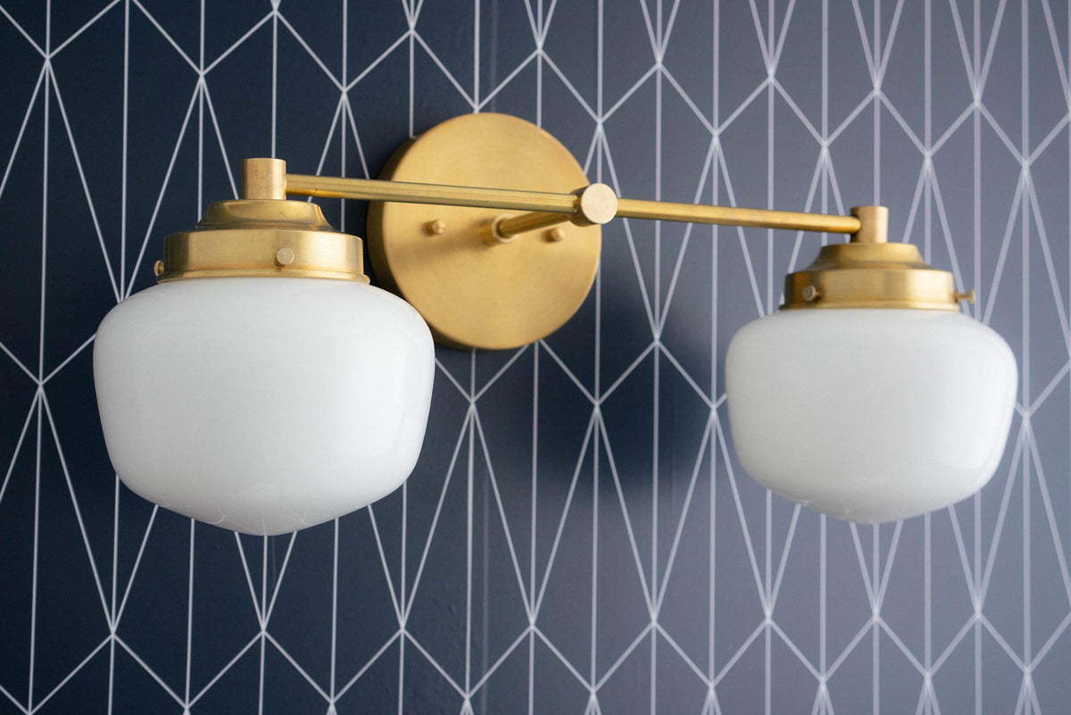 Raw brass two-bulb vanity light with opal globe shades and round wall mount against geometric dark blue wallpaper. - VANITY LIGHT MODEL NO. 5799
