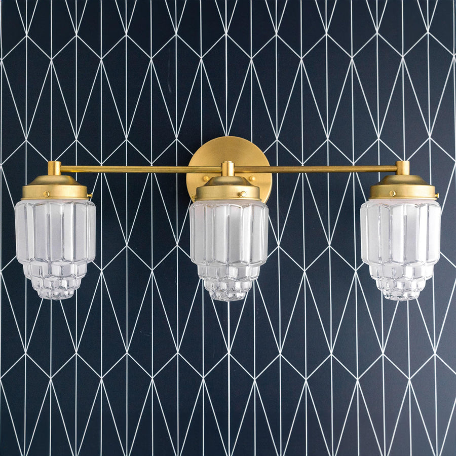 Art Deco vanity light with three clear globe shades and raw brass finish against geometric black wall. - VANITY LIGHT MODEL NO. 3918