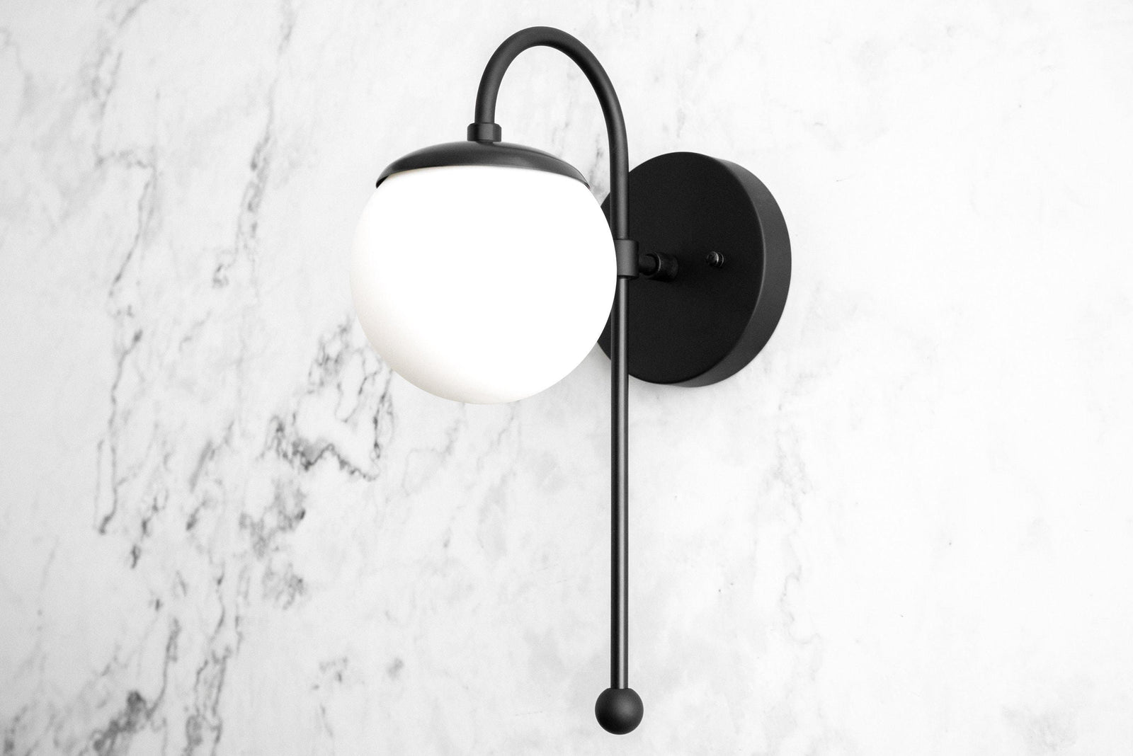 Black metal wall sconce with curved arm and frosted white globe shade on marble background. - SCONCE MODEL NO. 1045