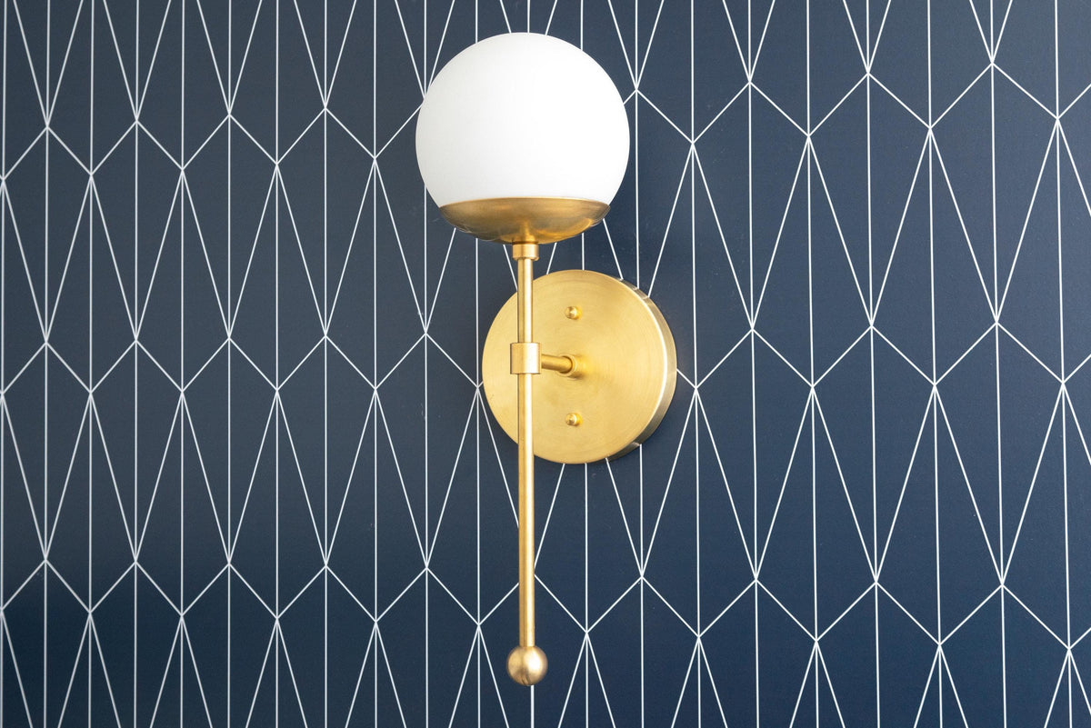 Art Deco wall sconce with a white globe shade and raw brass finish against dark geometric wallpaper. - SCONCE MODEL NO. 1048