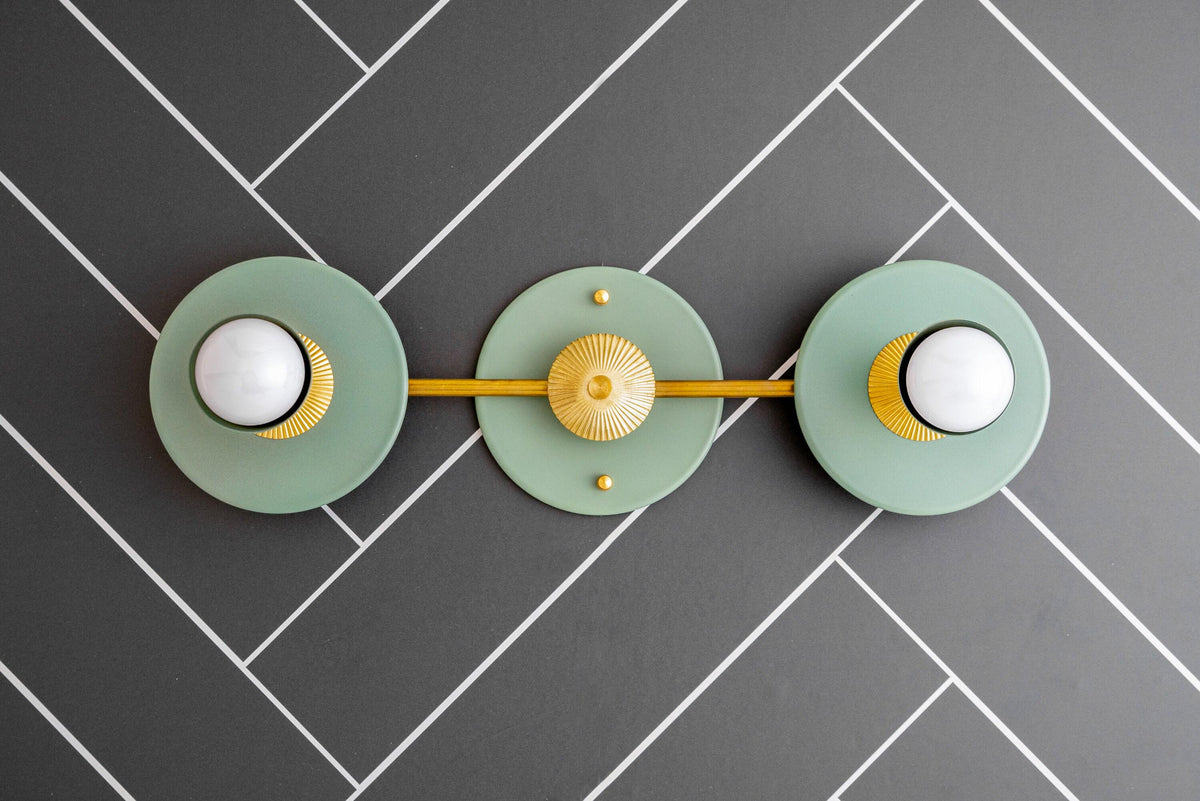 Raw brass vanity light with two green circular backplates and white bare bulbs on a gray herringbone tile wall. - VANITY LIGHT MODEL NO. 4473
