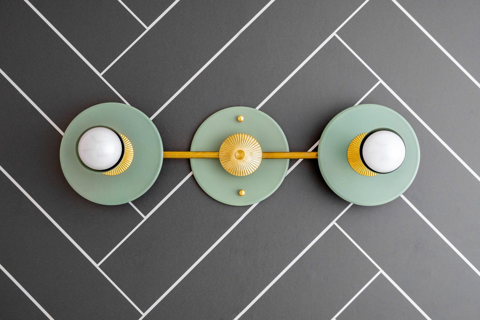 Raw brass vanity light with two green circular backplates and white bare bulbs on a gray herringbone tile wall. - VANITY LIGHT MODEL NO. 4473