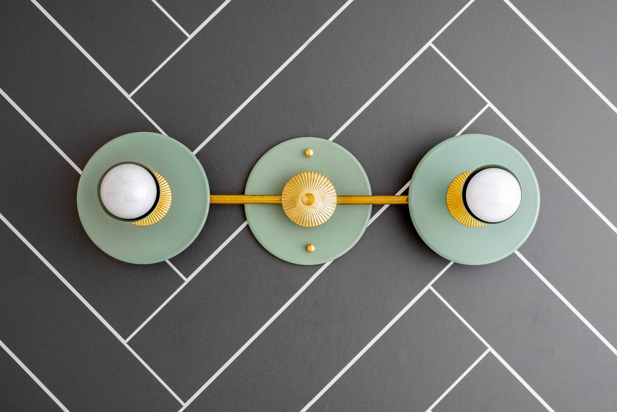 Raw brass vanity light with two green circular backplates and white bare bulbs on a gray herringbone tile wall. - VANITY LIGHT MODEL NO. 4473