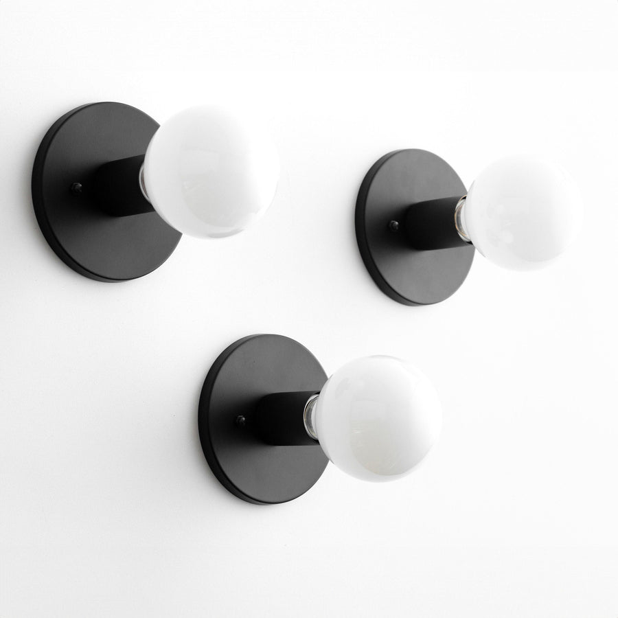 Set of three black flush-mount wall sconces with round backplates and exposed white globe bulbs. - SCONCE MODEL NO. 2057
